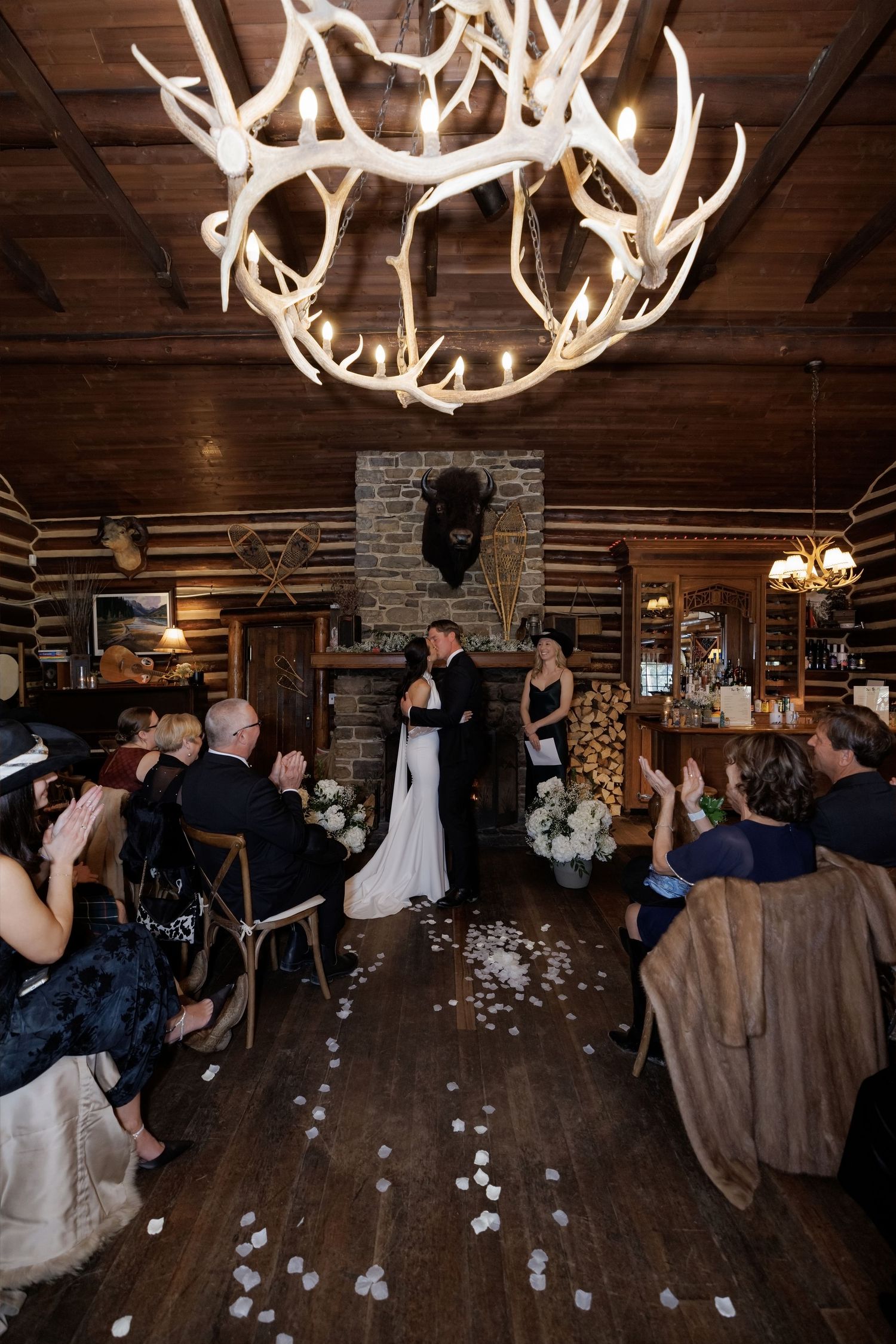 Couple shares first kiss at rustic log cabin wedding ceremony under antler chandelier with floral aisle.