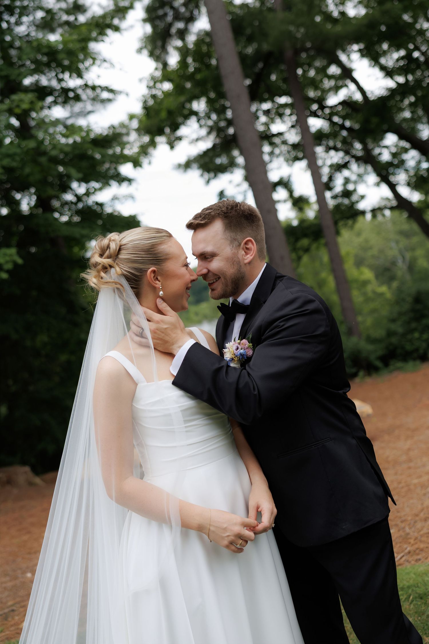 A bride and groom share a romantic moment outdoors, surrounded by lush green trees, as he tenderly holds her face.