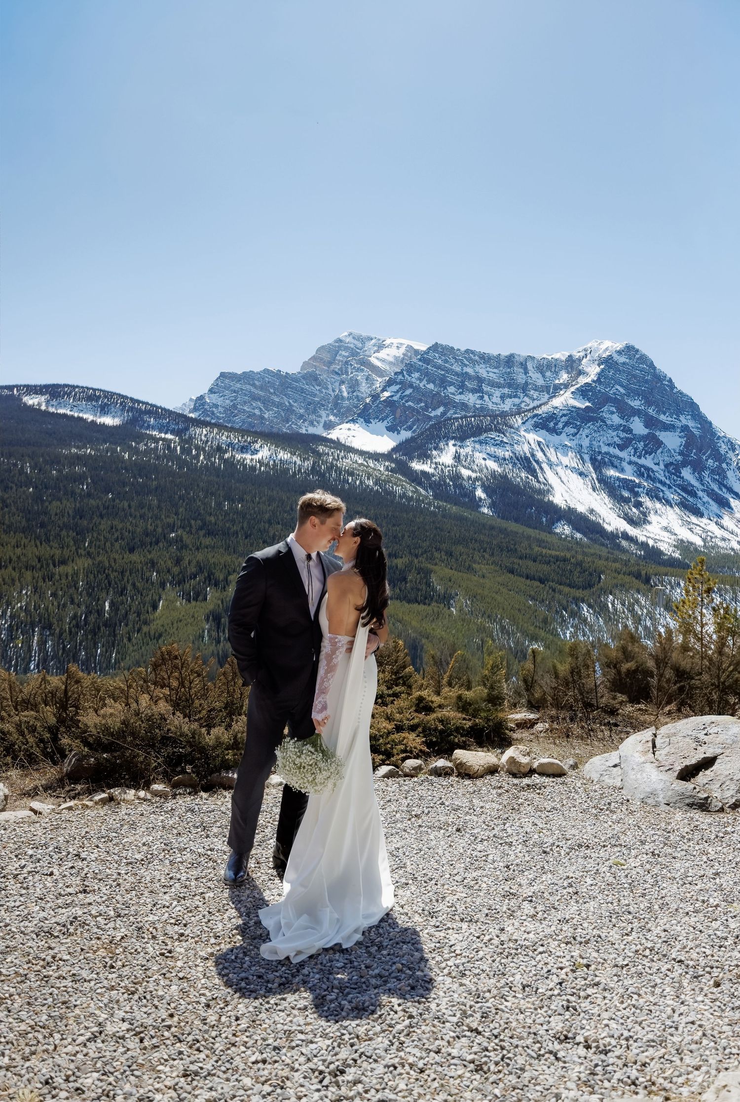 Bride and groom kissing in front of snow-capped mountain backdrop on rocky terrain during outdoor wedding ceremony.