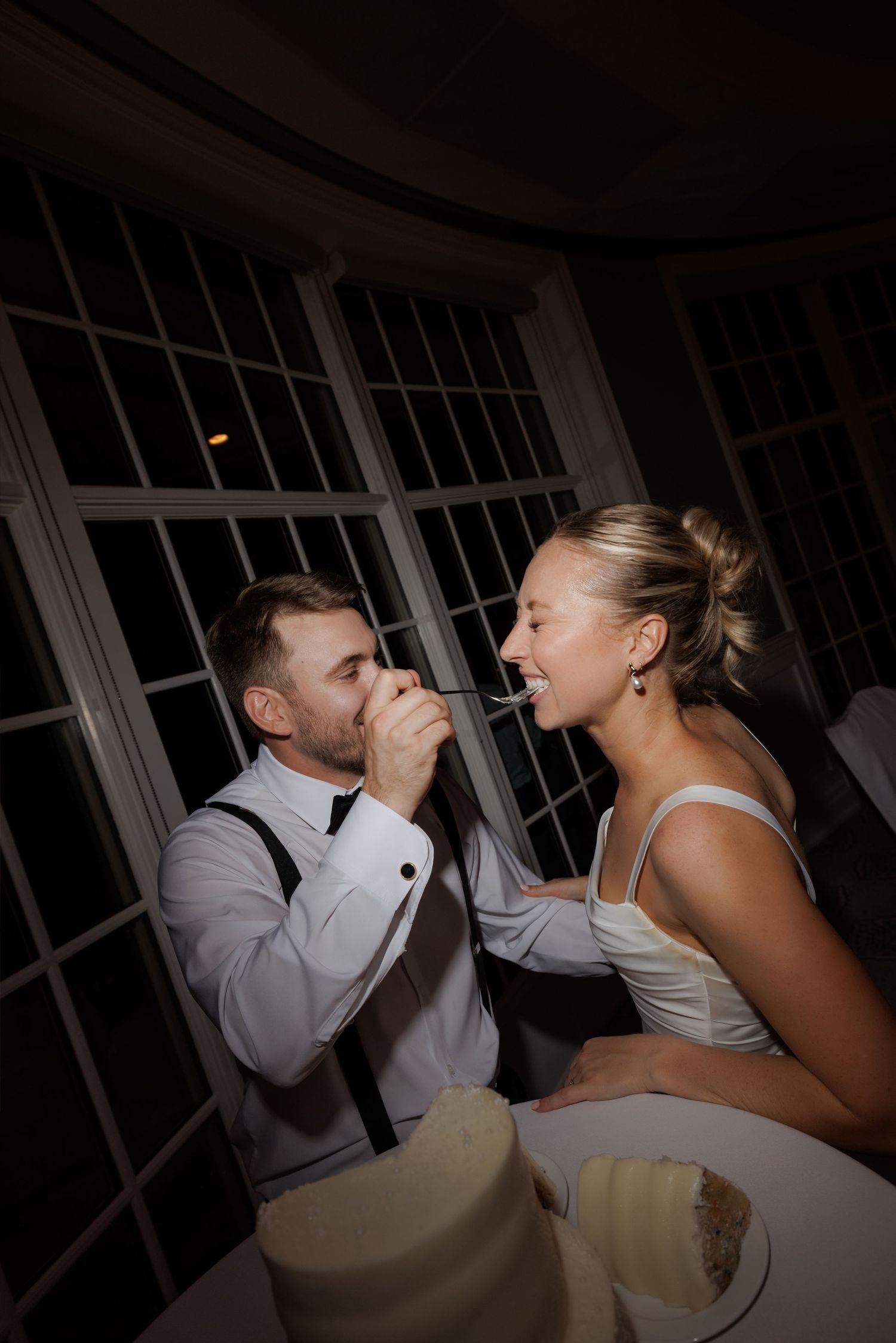 Bride and groom feed each other wedding cake at elegant indoor reception celebration.