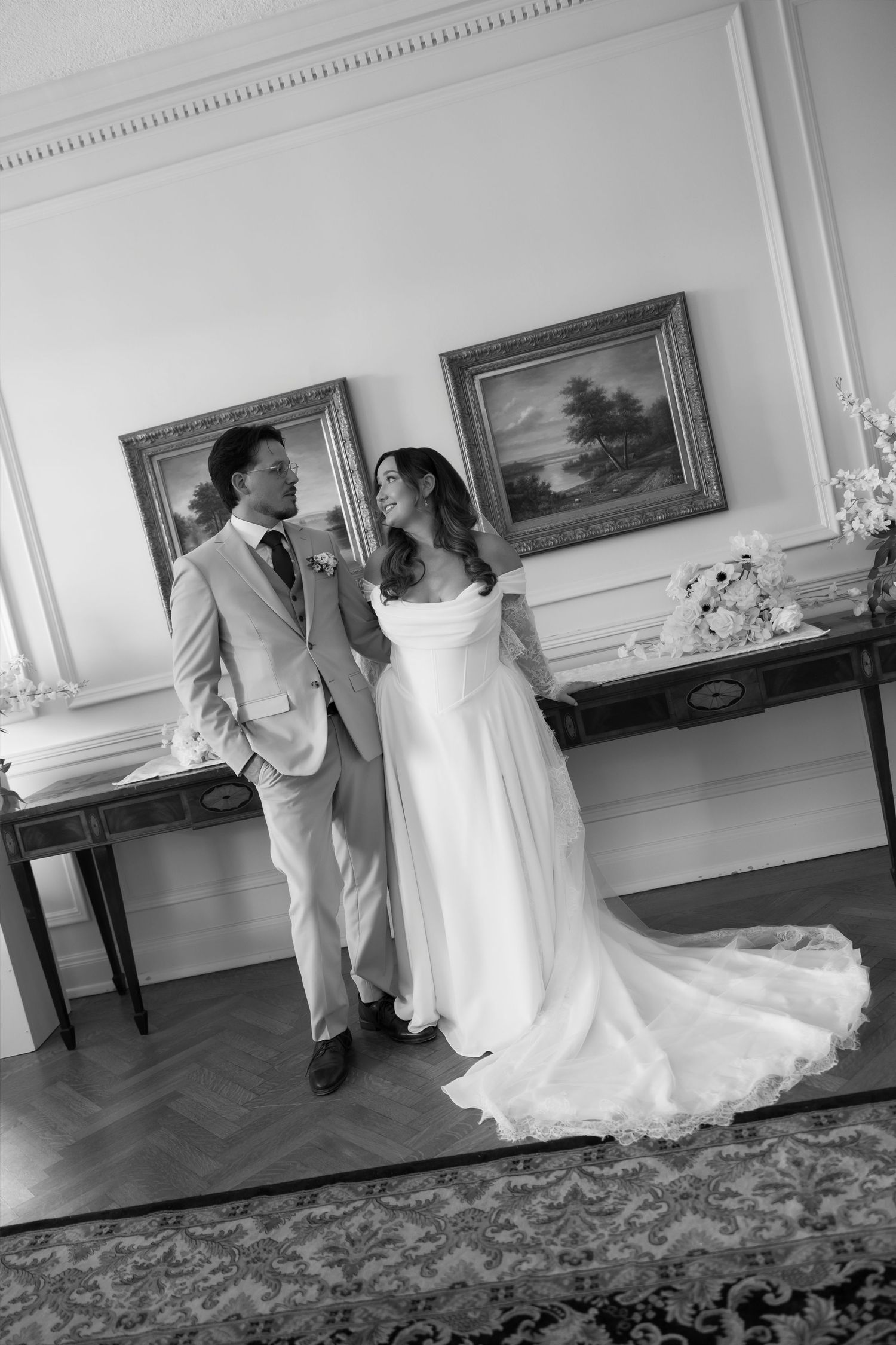 Black and white photo of a bride and groom sharing a romantic moment in an elegant room with framed artwork.