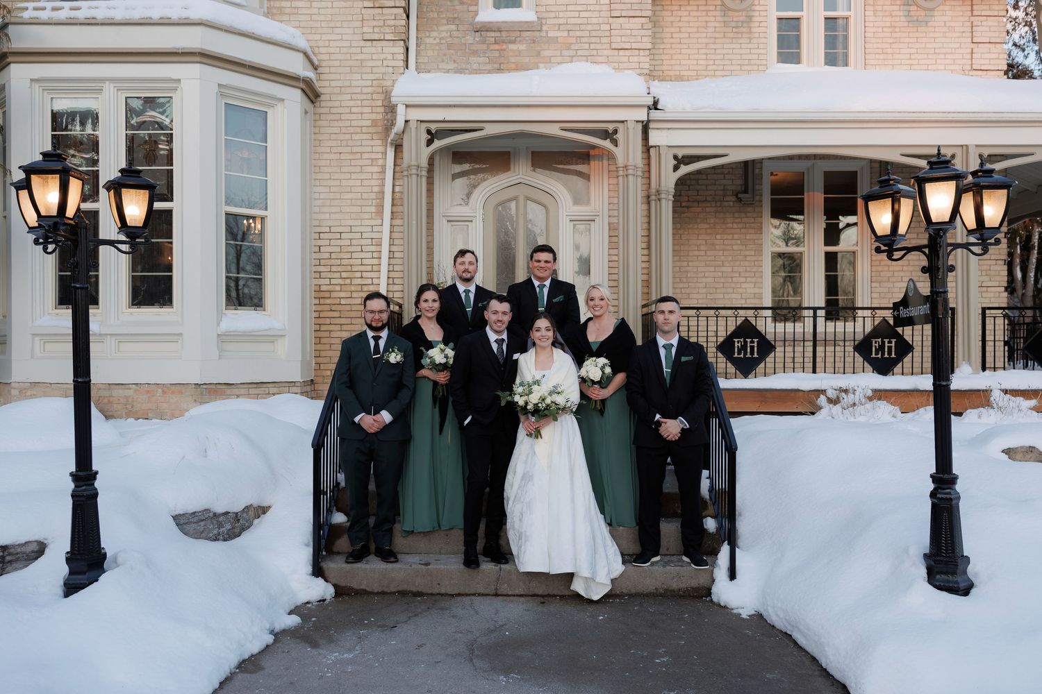 Wedding party poses in green and black attire outside a snow-covered historic venue with lamp posts.