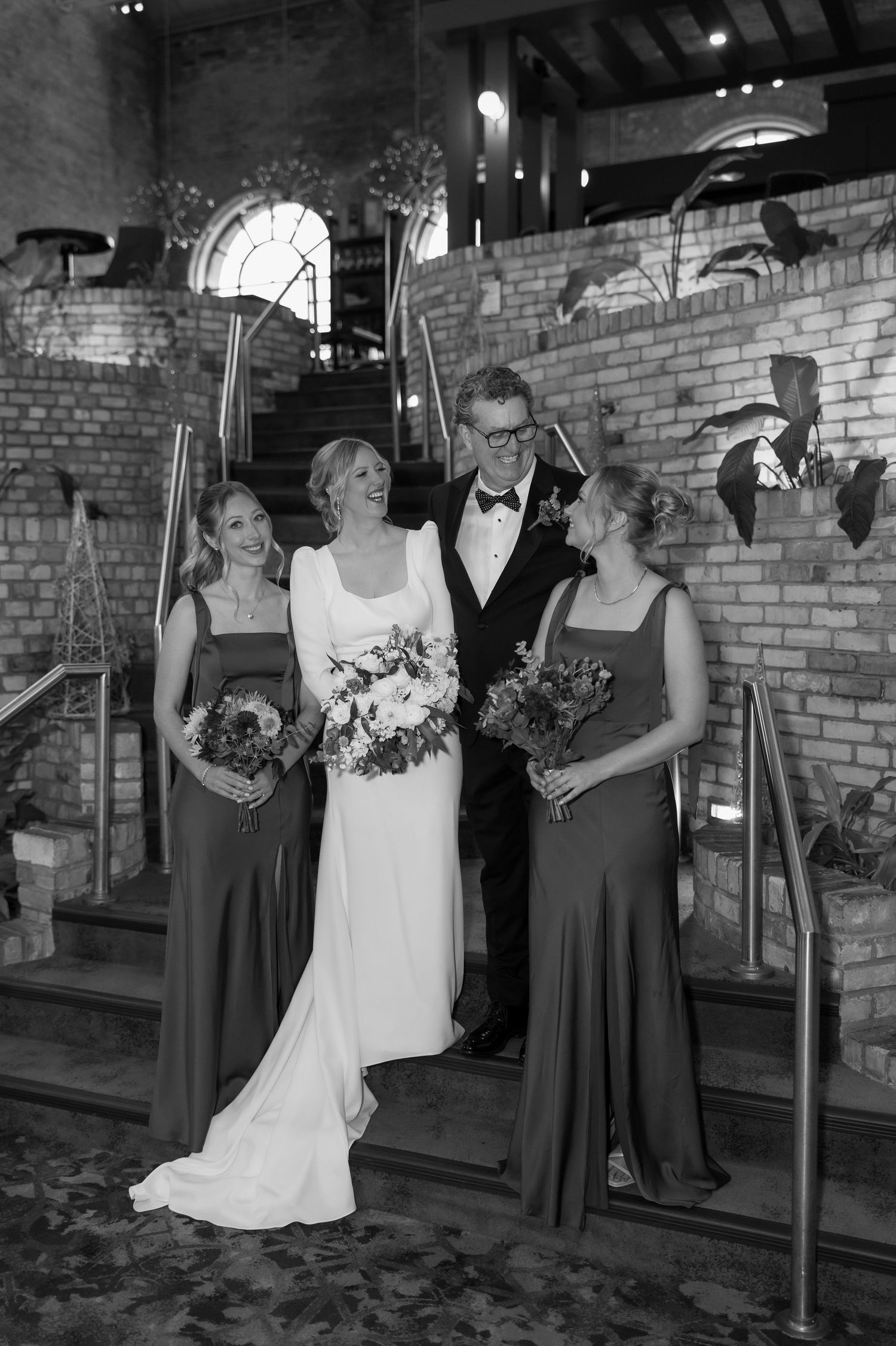 Wedding party poses against brick wall with bridesmaids in dark dresses and bride in white holding bouquets.