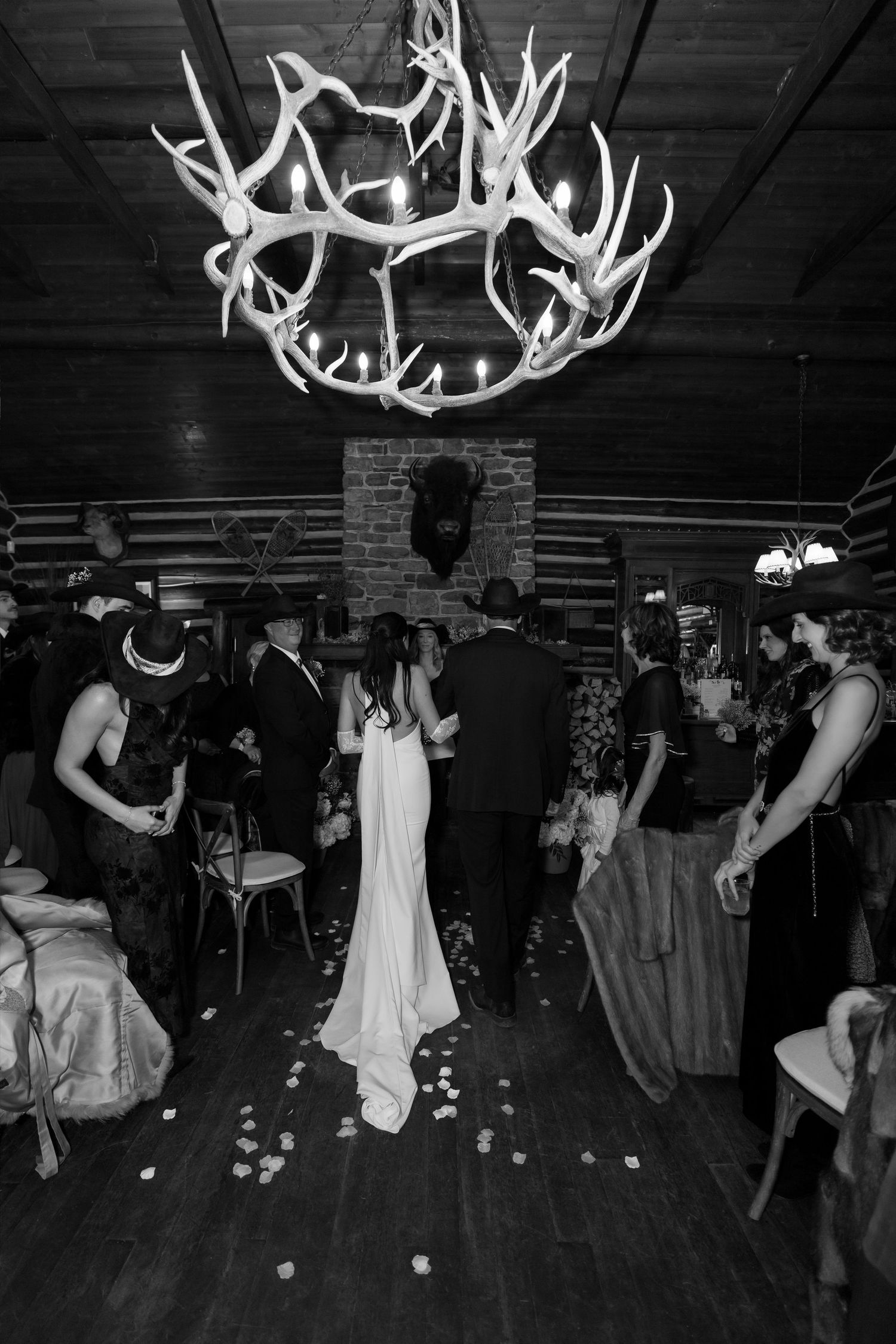 Black and white photo of bride walking down aisle under antler chandelier in rustic log cabin wedding venue.