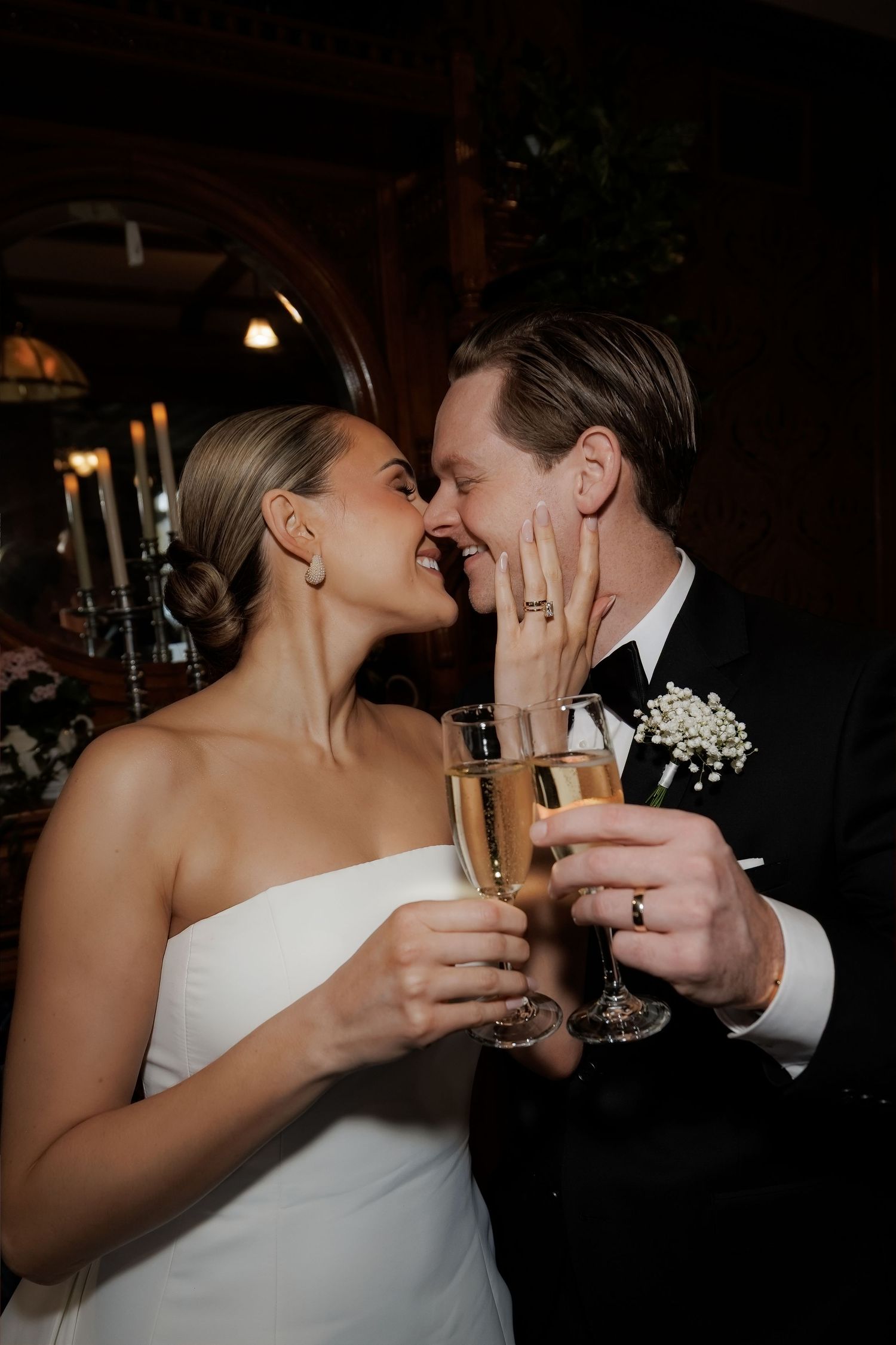 Bride and groom toasting with champagne flutes, touching noses and smiling, wearing wedding attire in a dimly lit venue.