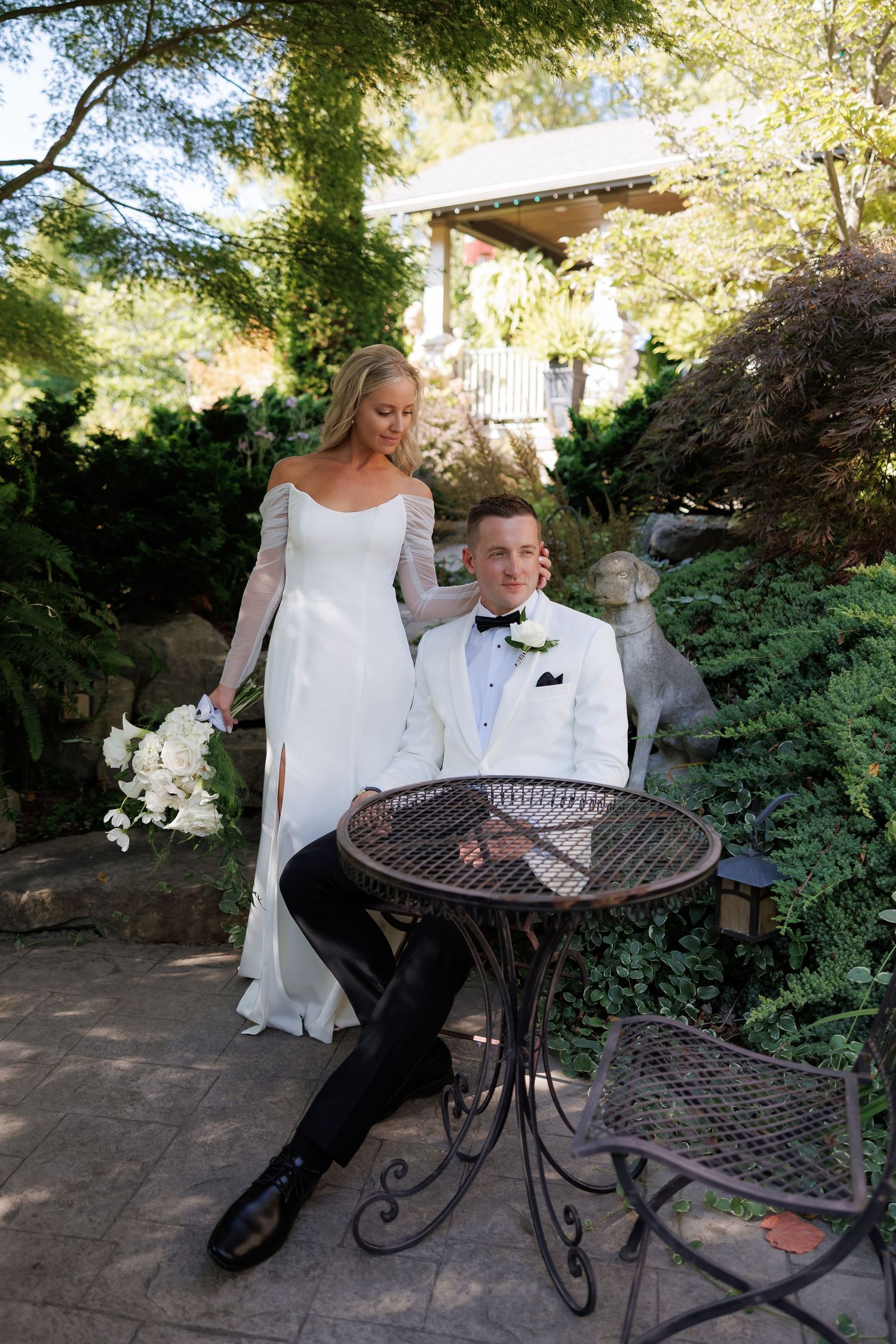 Bride in white gown holding bouquet stands beside seated groom in white tuxedo at garden wedding venue.