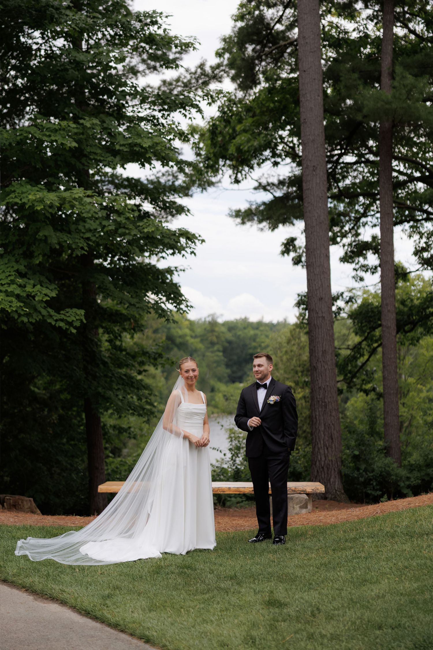 Bride in white gown with long veil and groom in black tuxedo standing on green lawn surrounded by tall trees.