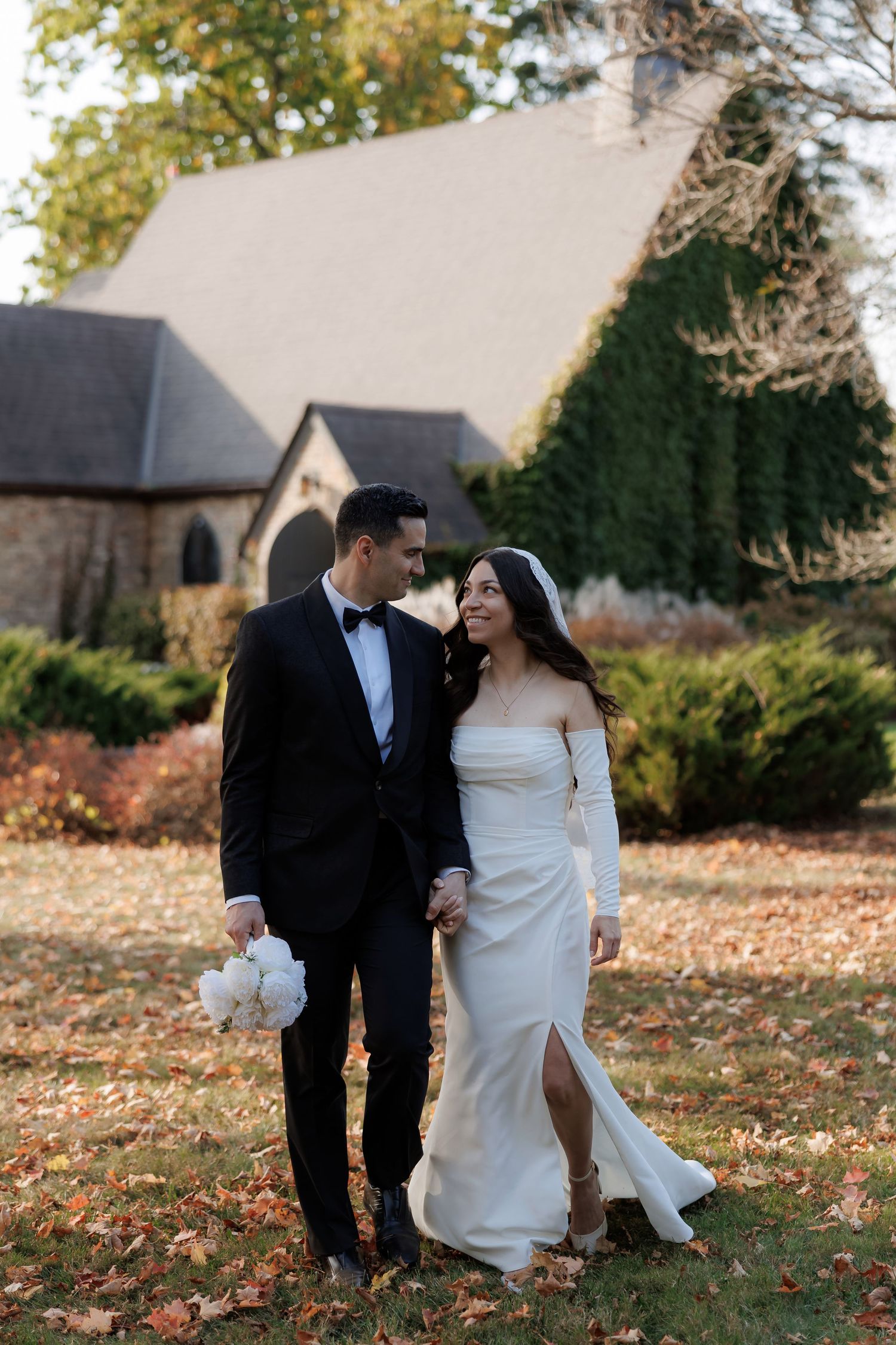 Romantic autumn wedding photos showing couple in formal attire walking through fallen leaves on church grounds.