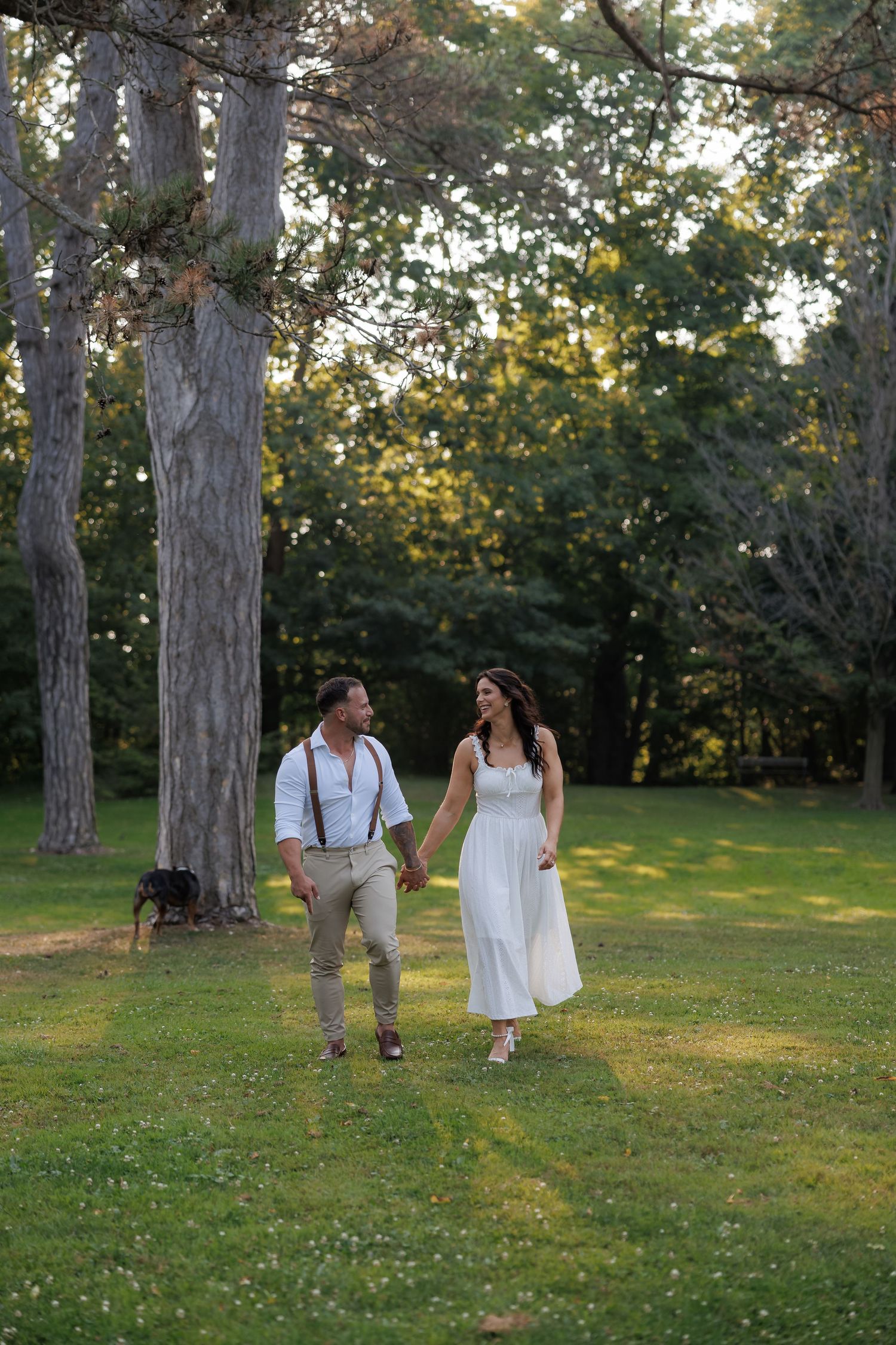 Couple walking through a sunlit park with tall pine trees.