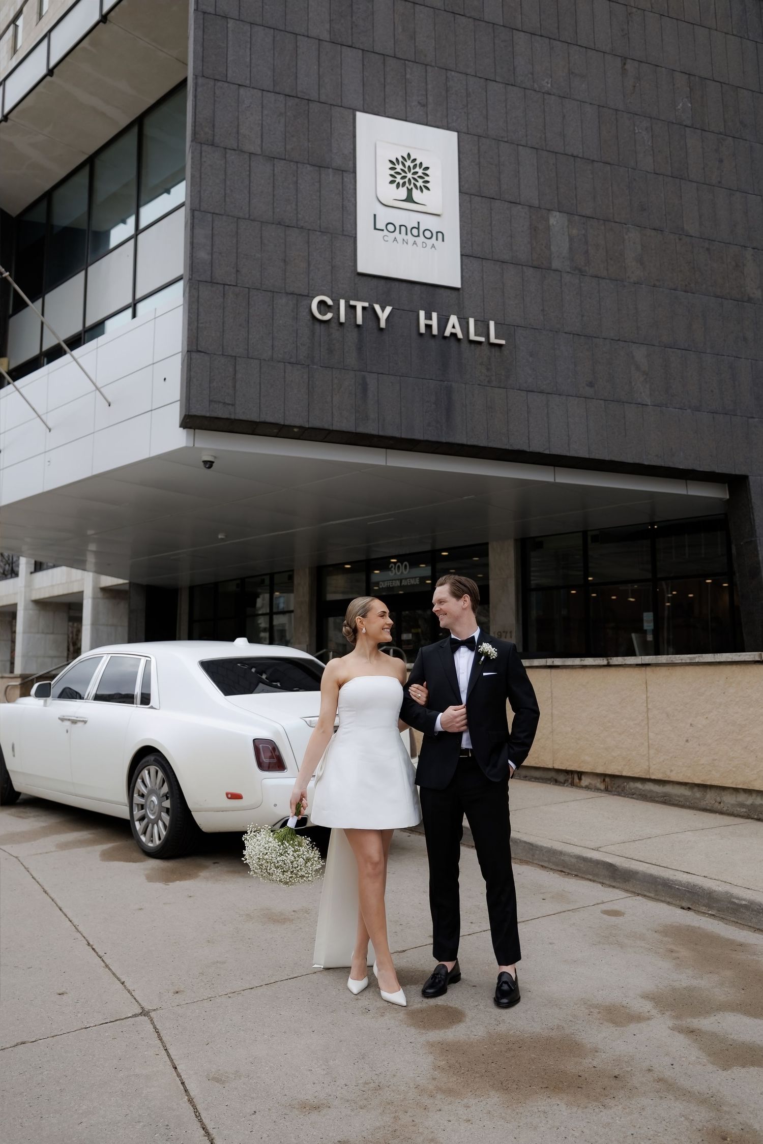 Bride in short white dress and groom in tuxedo pose before London City Hall with white Rolls-Royce.