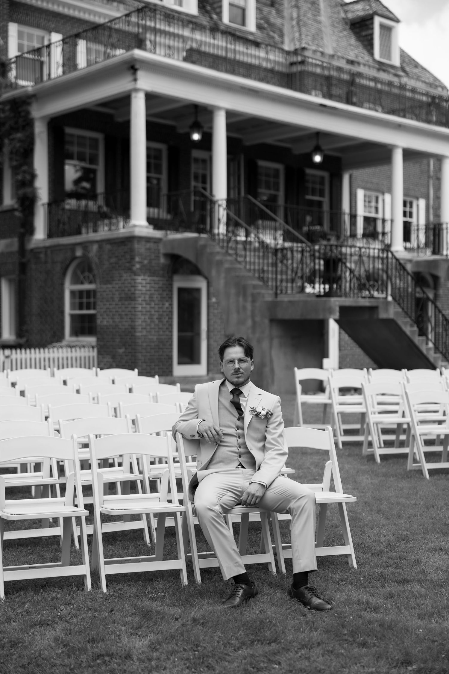 Groom in light suit sits on white folding chair at outdoor wedding ceremony setup before historic brick building.
