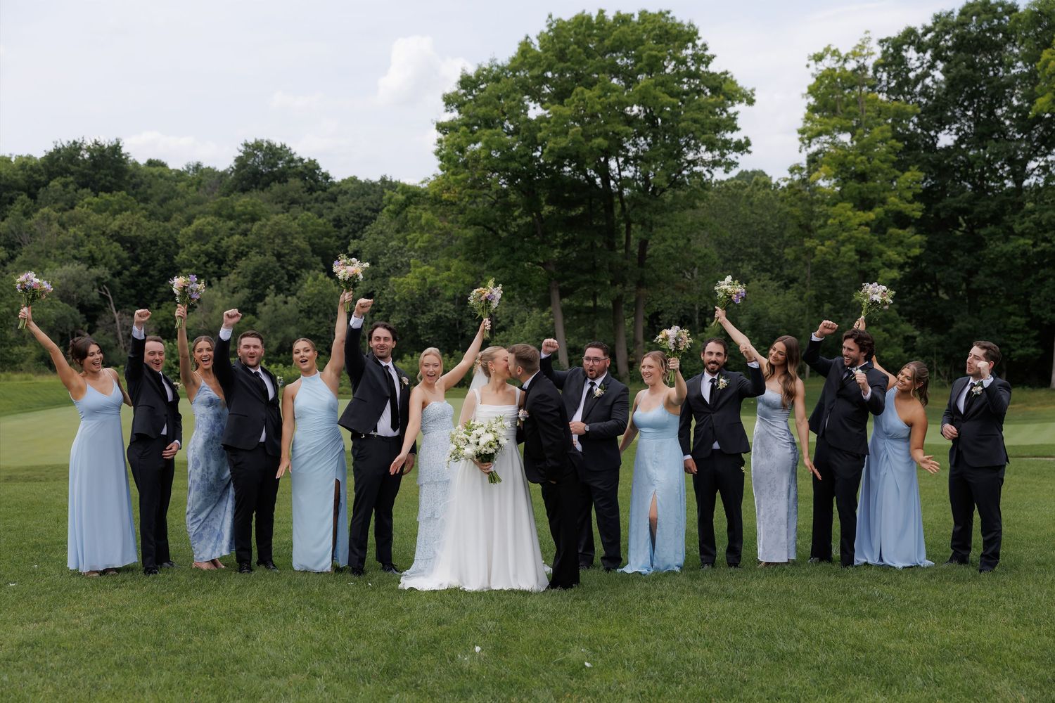 Wedding party in blue dresses and black tuxedos cheering with bouquets raised on green lawn.