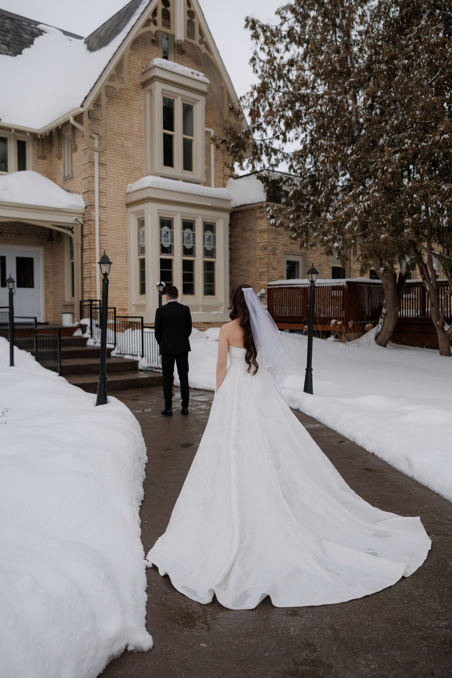 Bride in white gown with long train walks toward groom outside snow-covered stone wedding venue in winter.