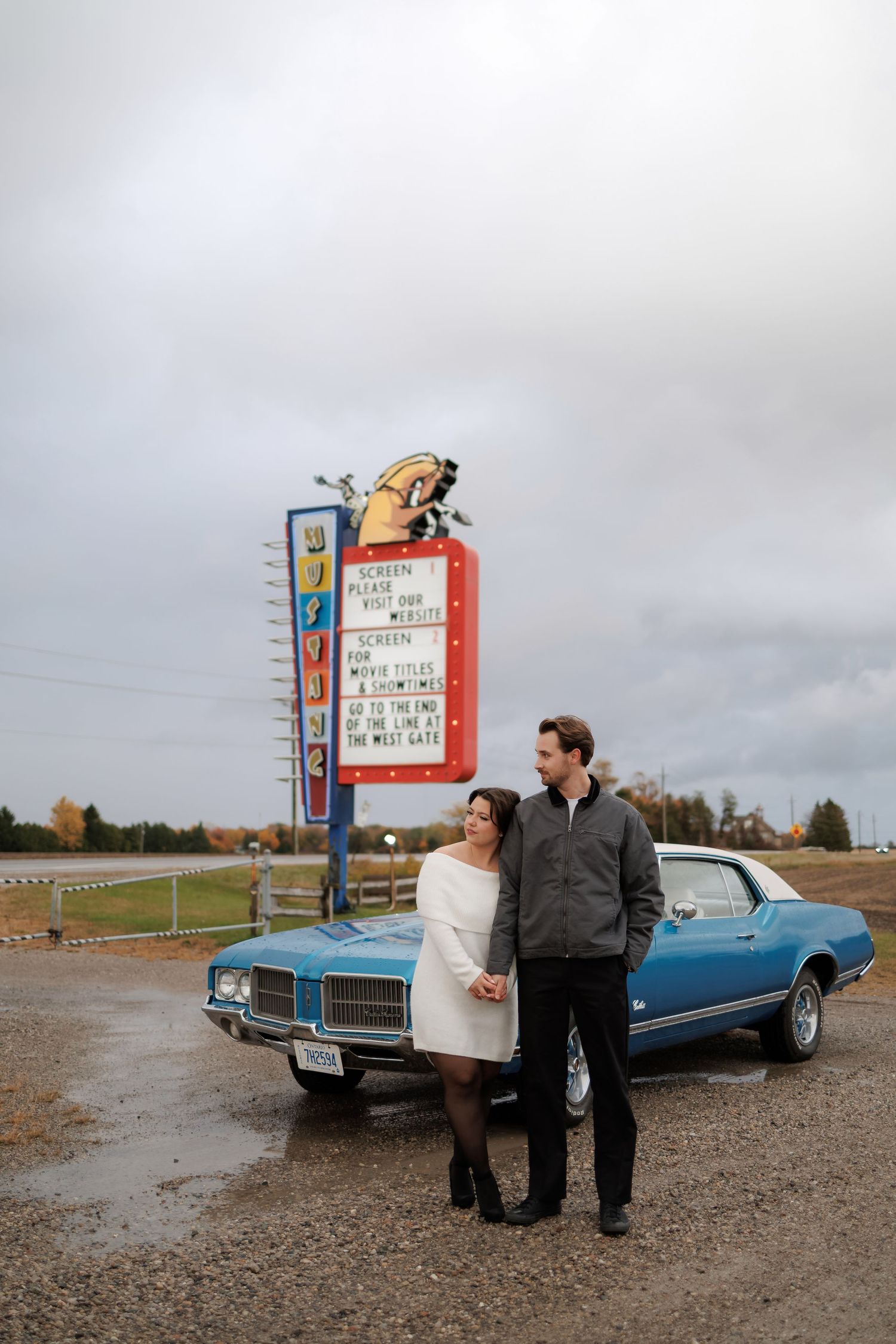 A couple stands near their classic blue car at a vintage drive-in theater with a retro marquee sign.
