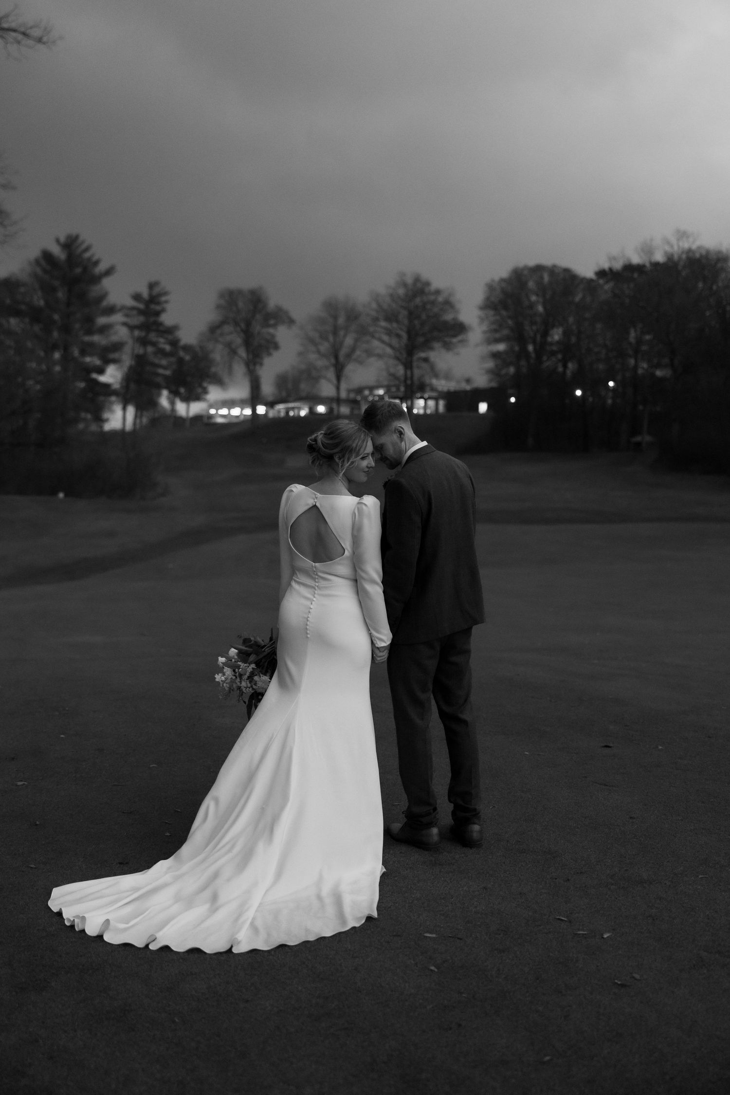A couple in wedding attire stands together on a golf course at dusk in a series of black and white photographs.