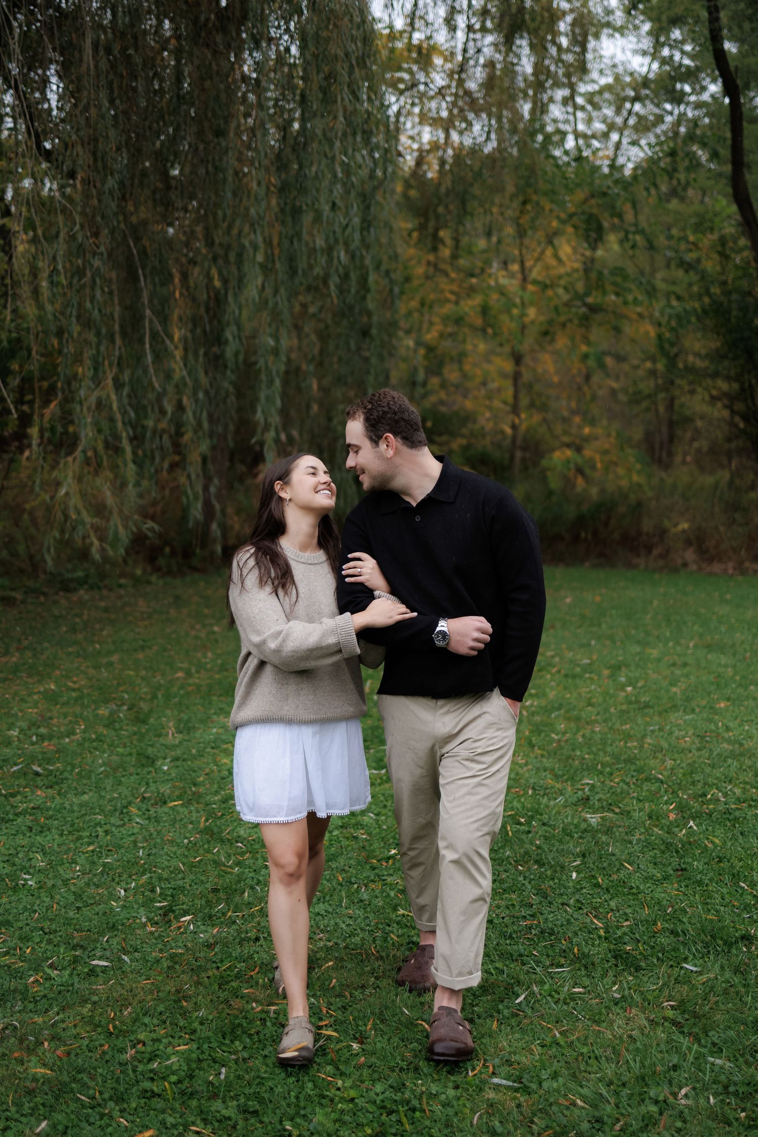 A couple shares a romantic moment while walking together on grass beneath autumn trees and hanging willow branches.