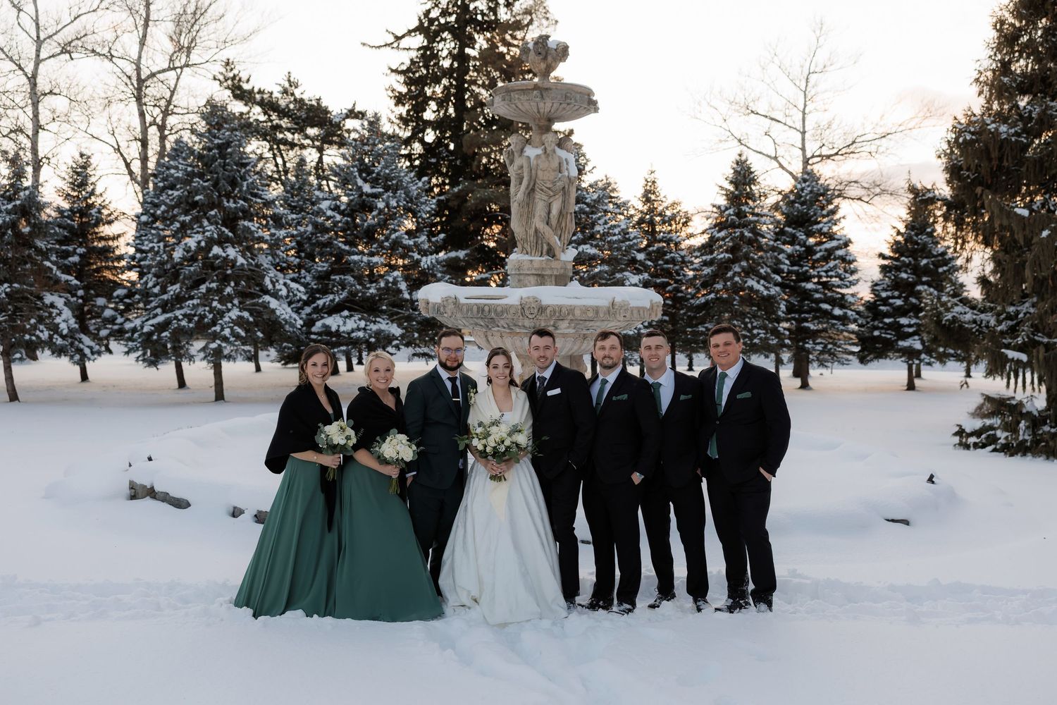 Wedding party poses in snow before a stone fountain, bridesmaids in green gowns and groomsmen in black suits.