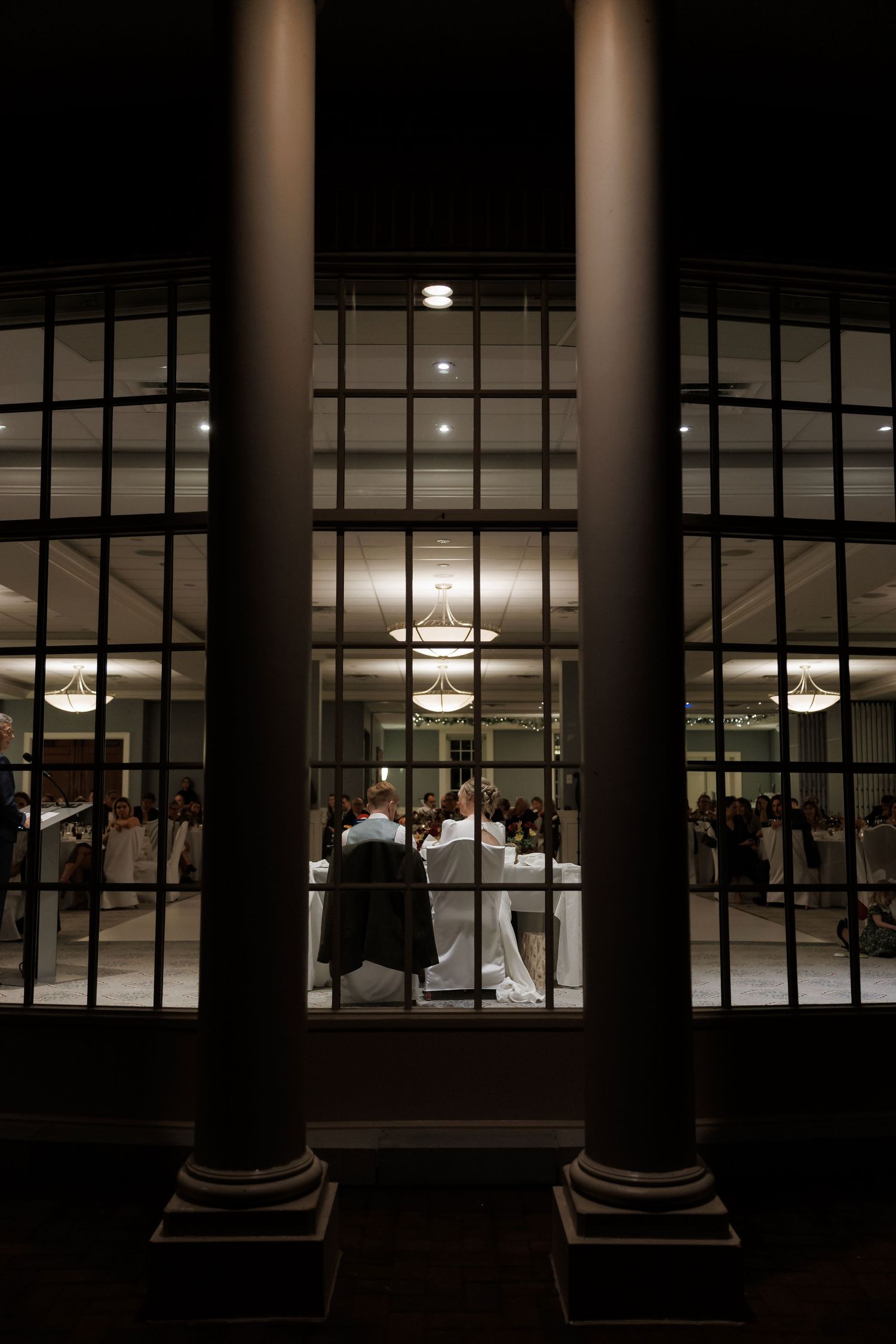 Wedding reception venue viewed through elegant columns at night with chandeliers glowing inside.