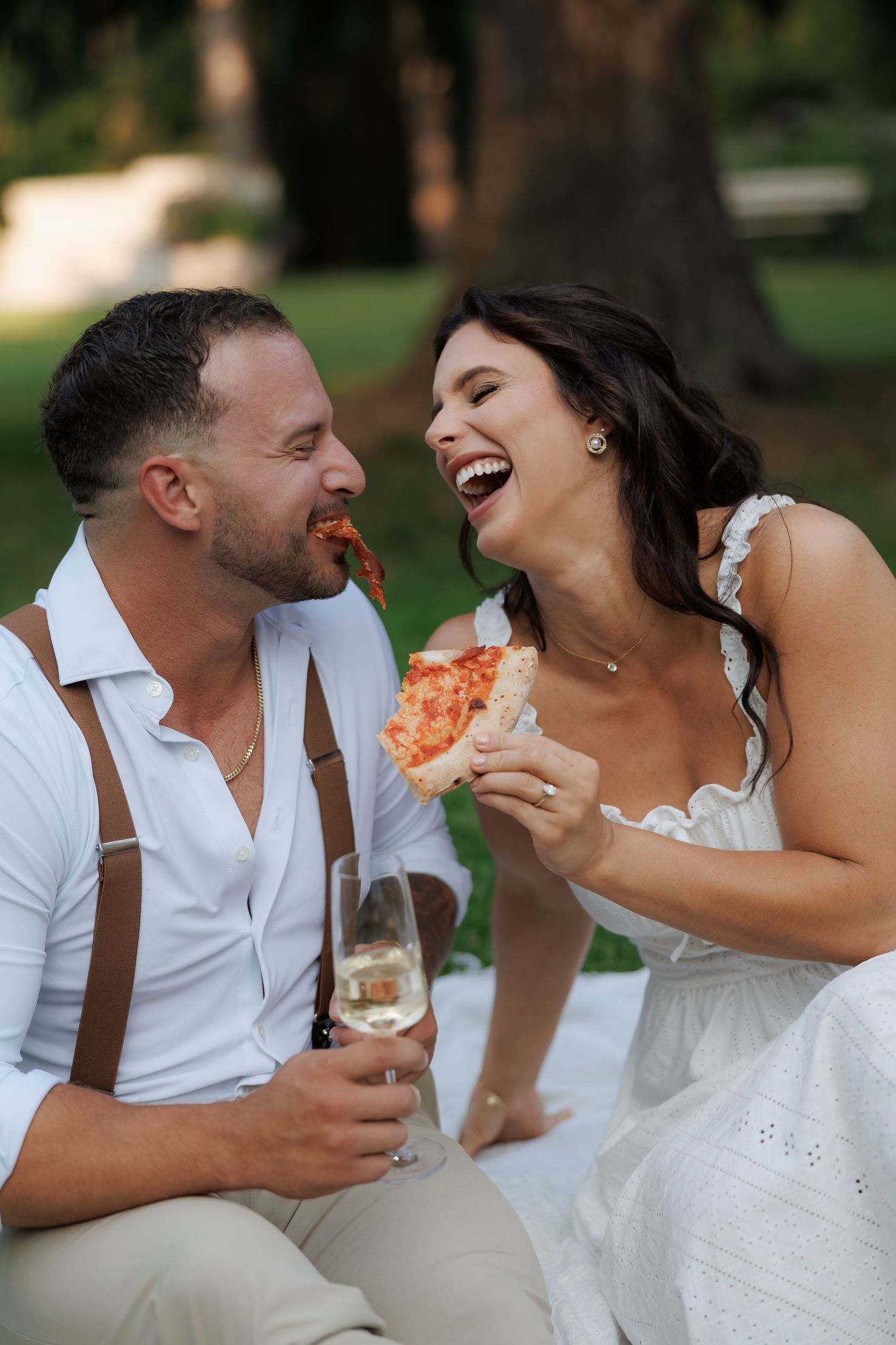 Candid moment of couple laughing while sharing pizza during outdoor picnic.