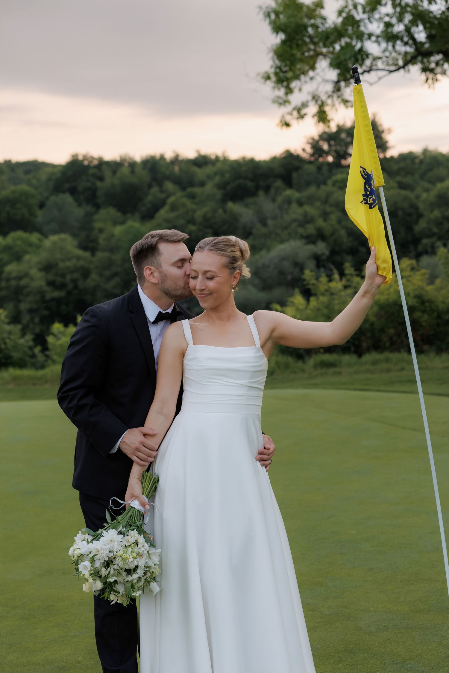 Groom kissing brides cheek as she holds a yellow golf flag on a golf course at sunset.