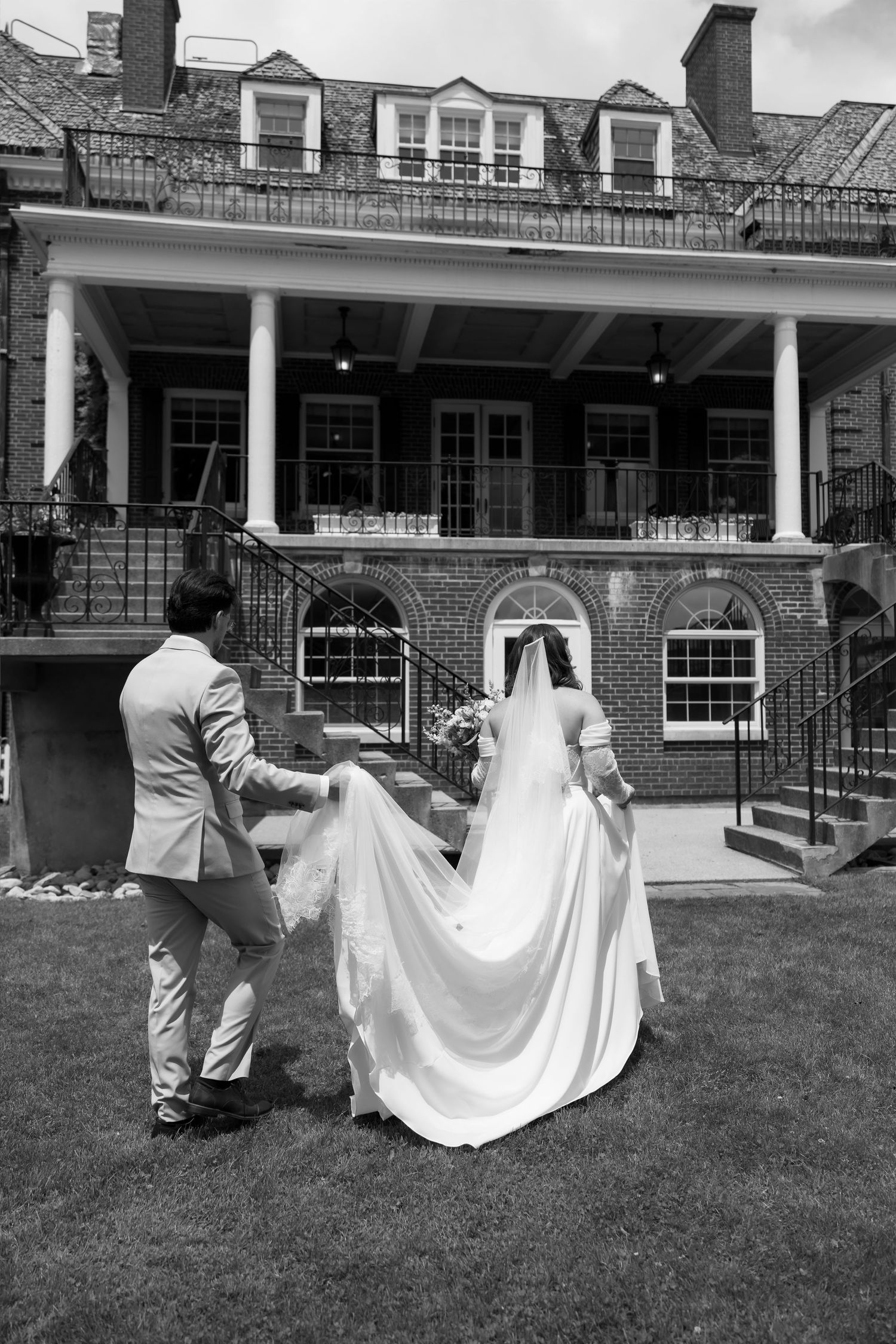 Black and white photo of a groom holding his bride's wedding dress train as they walk toward an elegant brick mansion.