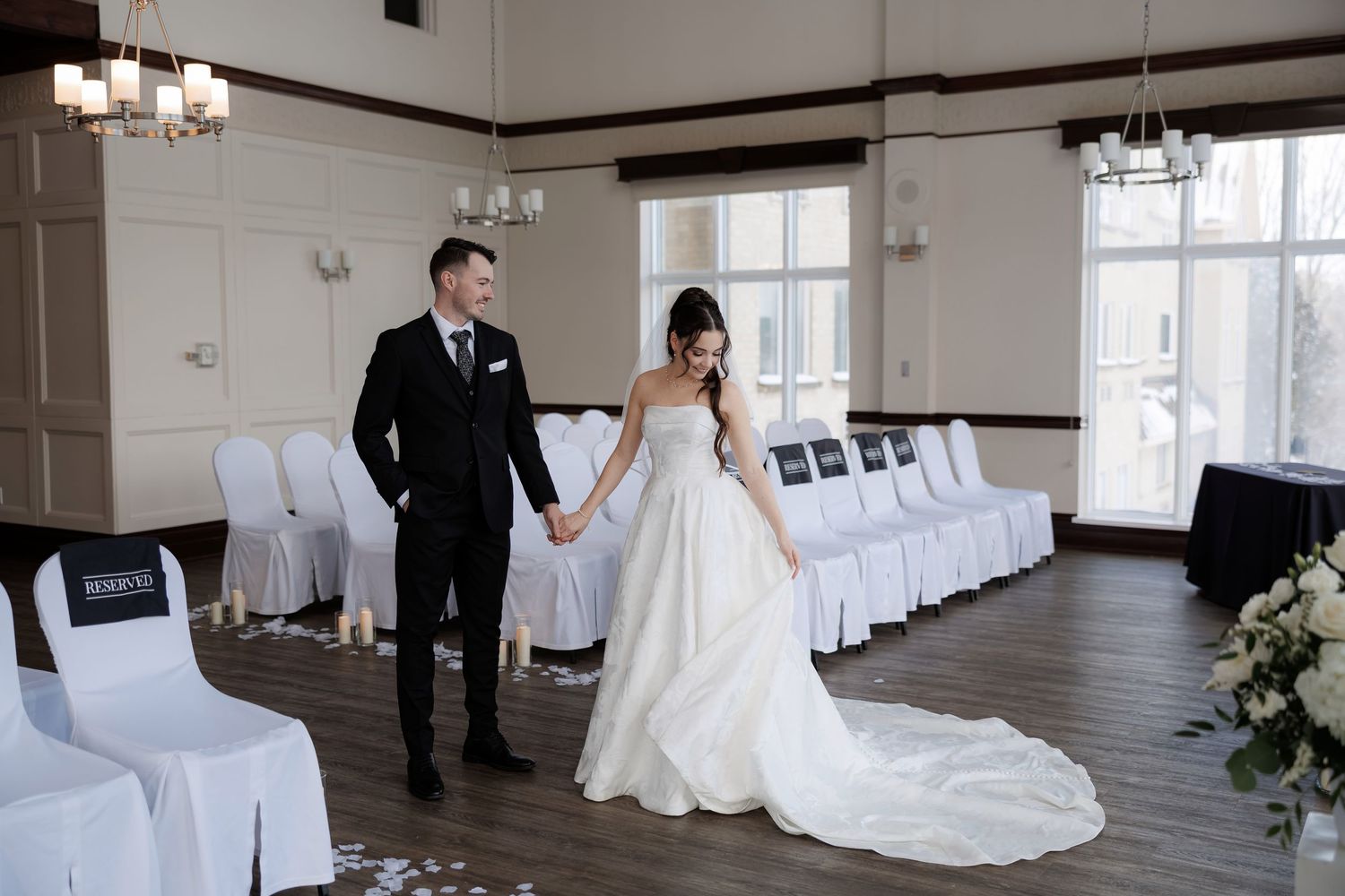 Bride and groom holding hands in elegant white wedding venue with draped chairs and chandelier lighting.
