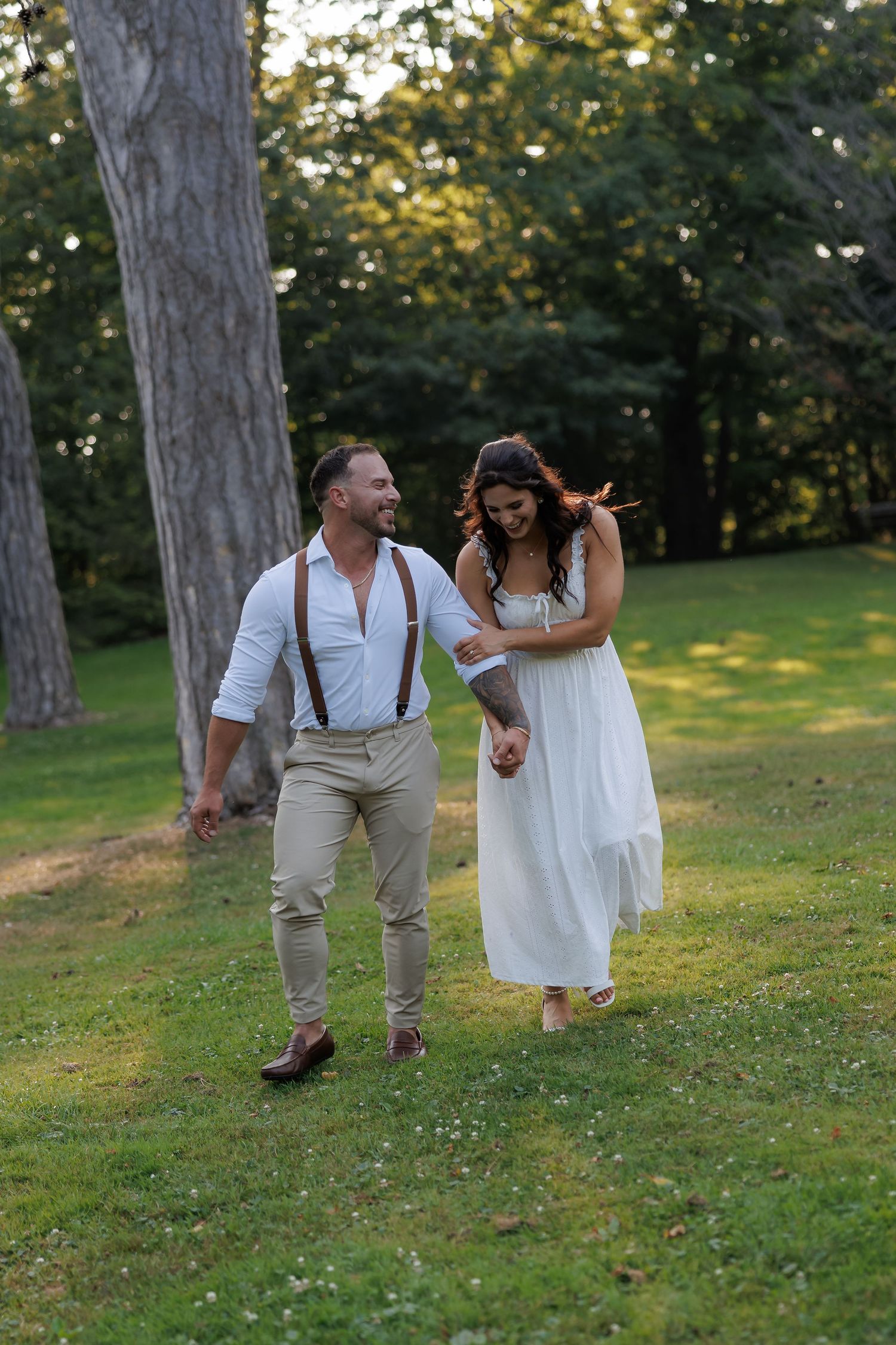 A couple in white and beige attire walk hand in hand through a grassy hillside with trees in the background at sunset.