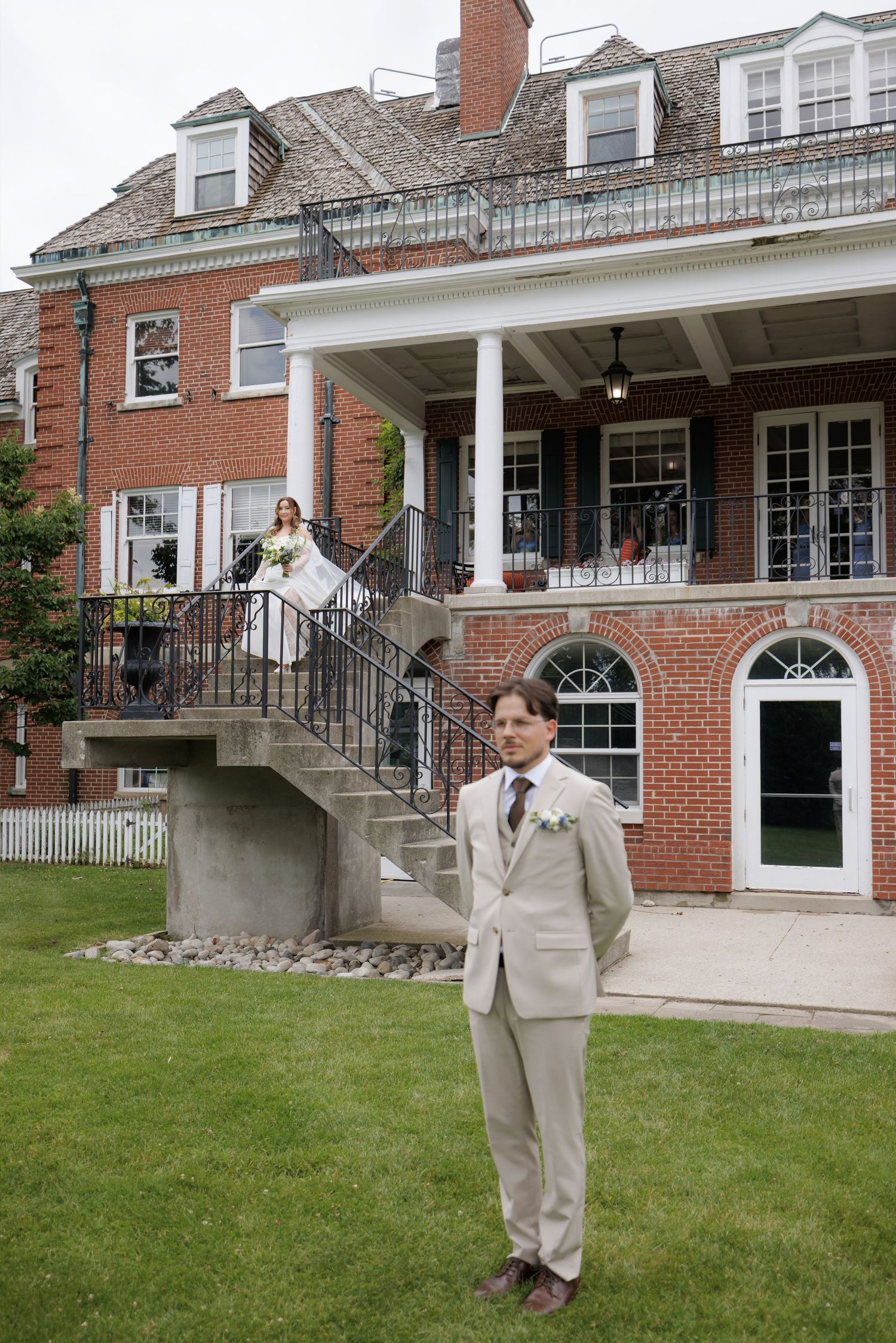 Groom in tan suit stands on lawn as bride descends grand staircase of brick wedding venue.