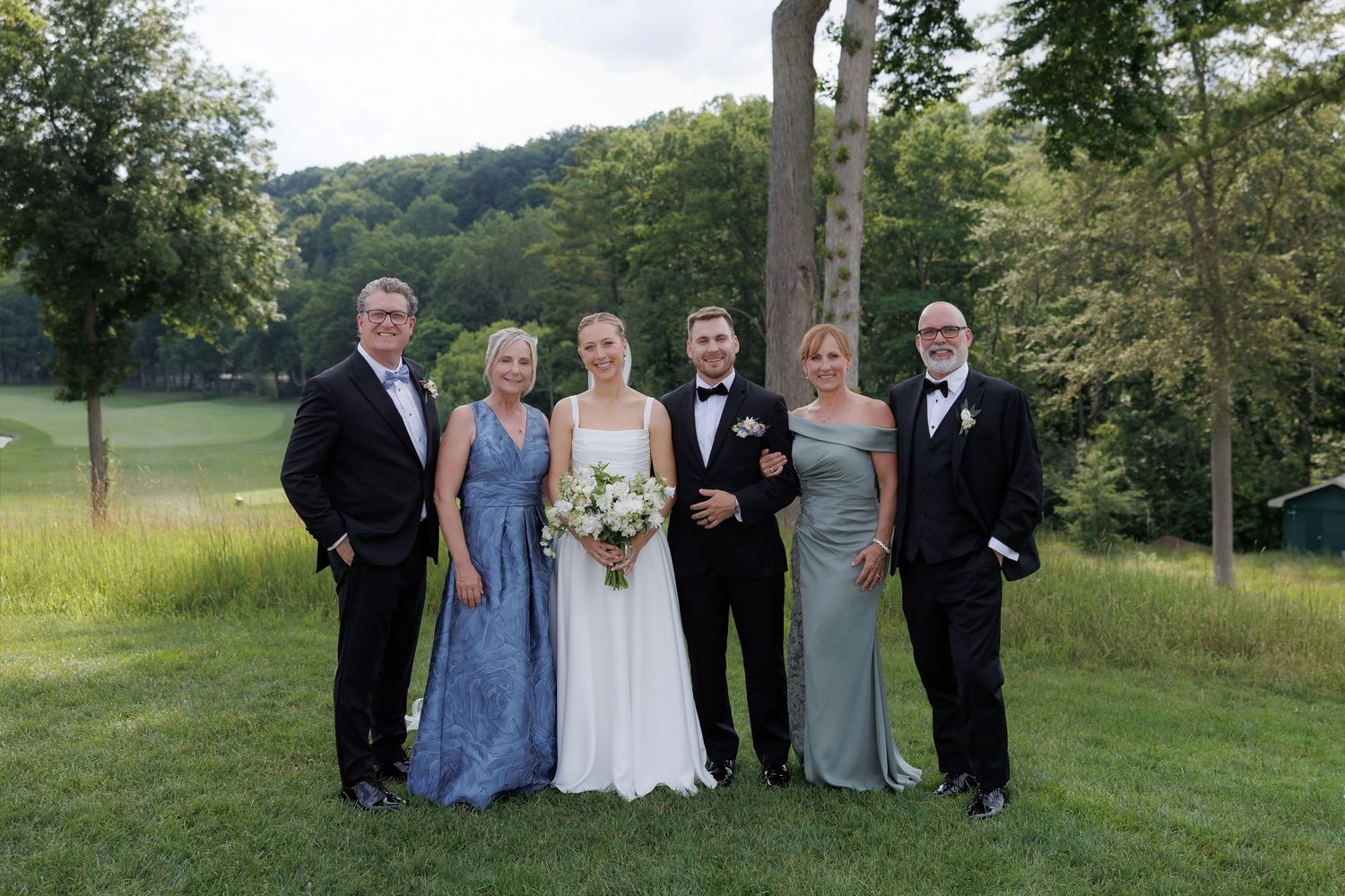 Wedding party of six posing outdoors on green lawn with wooded hillside backdrop, bride in white gown holding bouquet.
