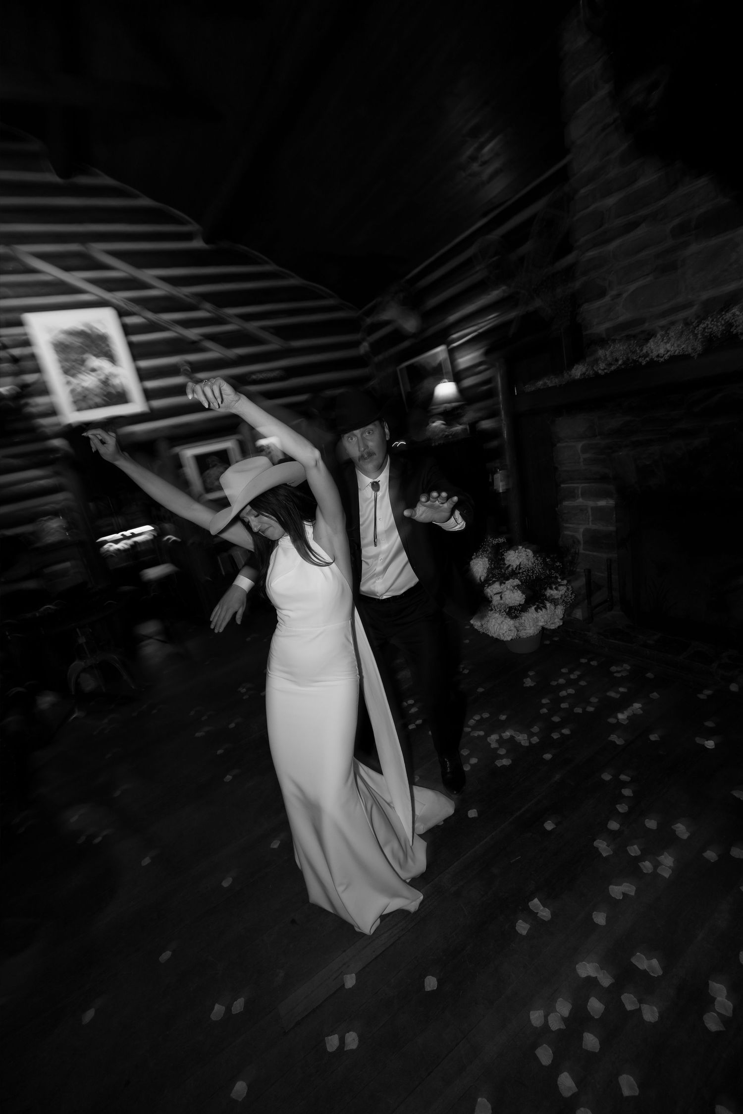 Black and white photo of bride in white gown and cowboy hat dancing with groom at rustic wedding reception.