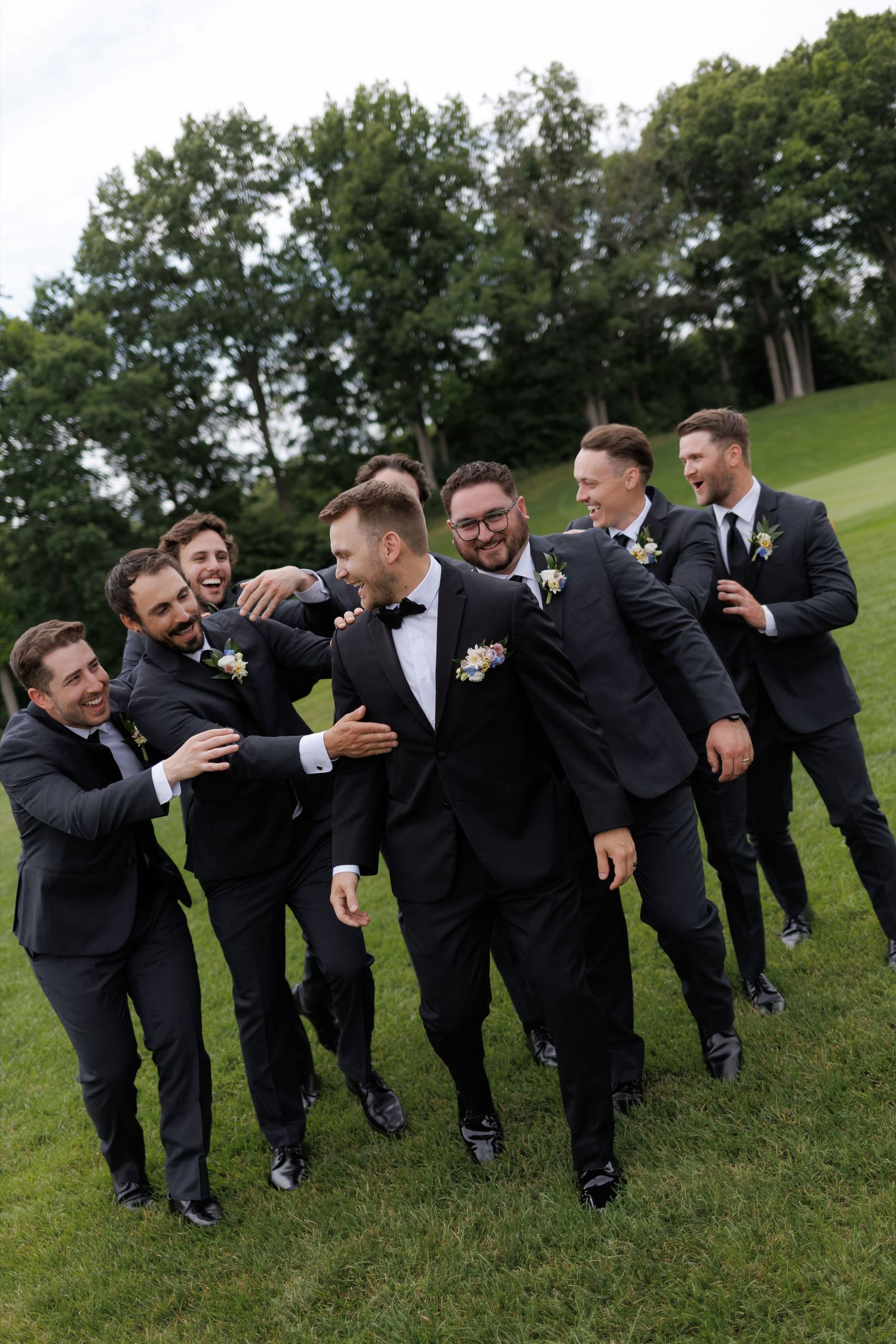 Groom laughing with groomsmen in black suits on a green lawn during wedding party photos.