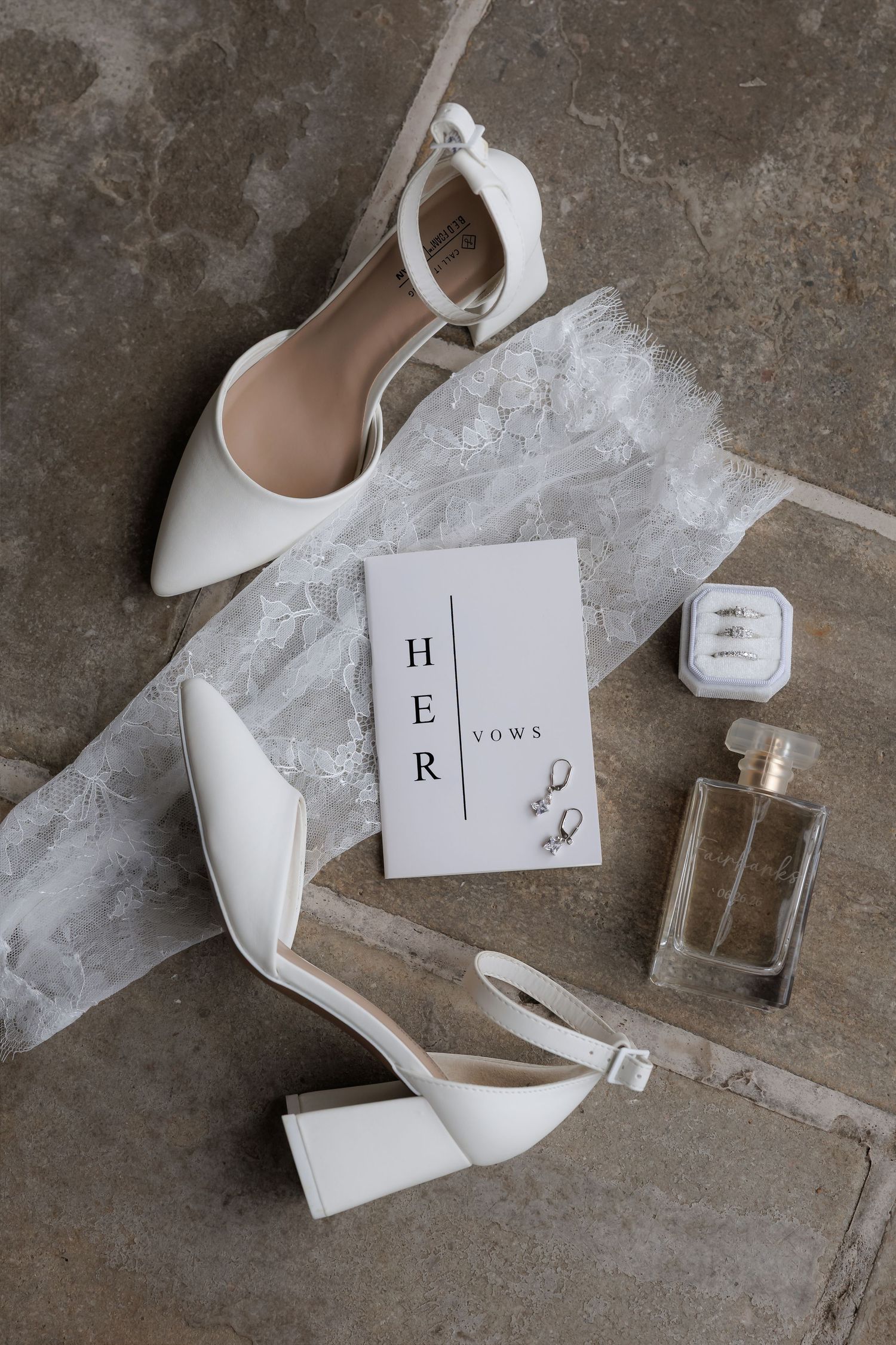 White bridal heels, vow booklet, pearl earrings, perfume bottle, and lace garter arranged on stone floor.