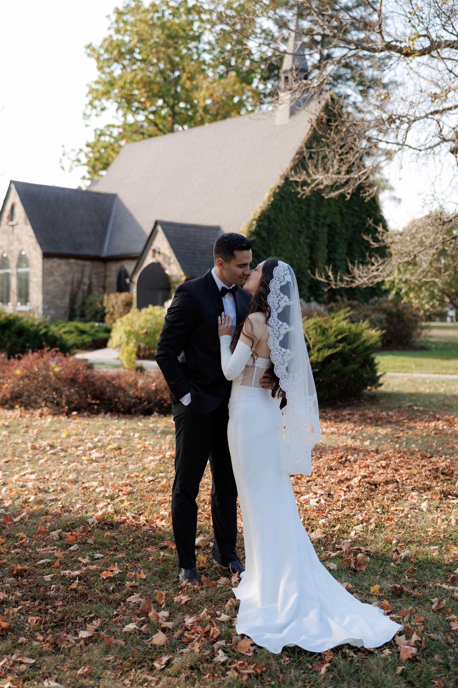 Romantic autumn wedding photos with couple embracing on leaf-covered lawn near stone church.
