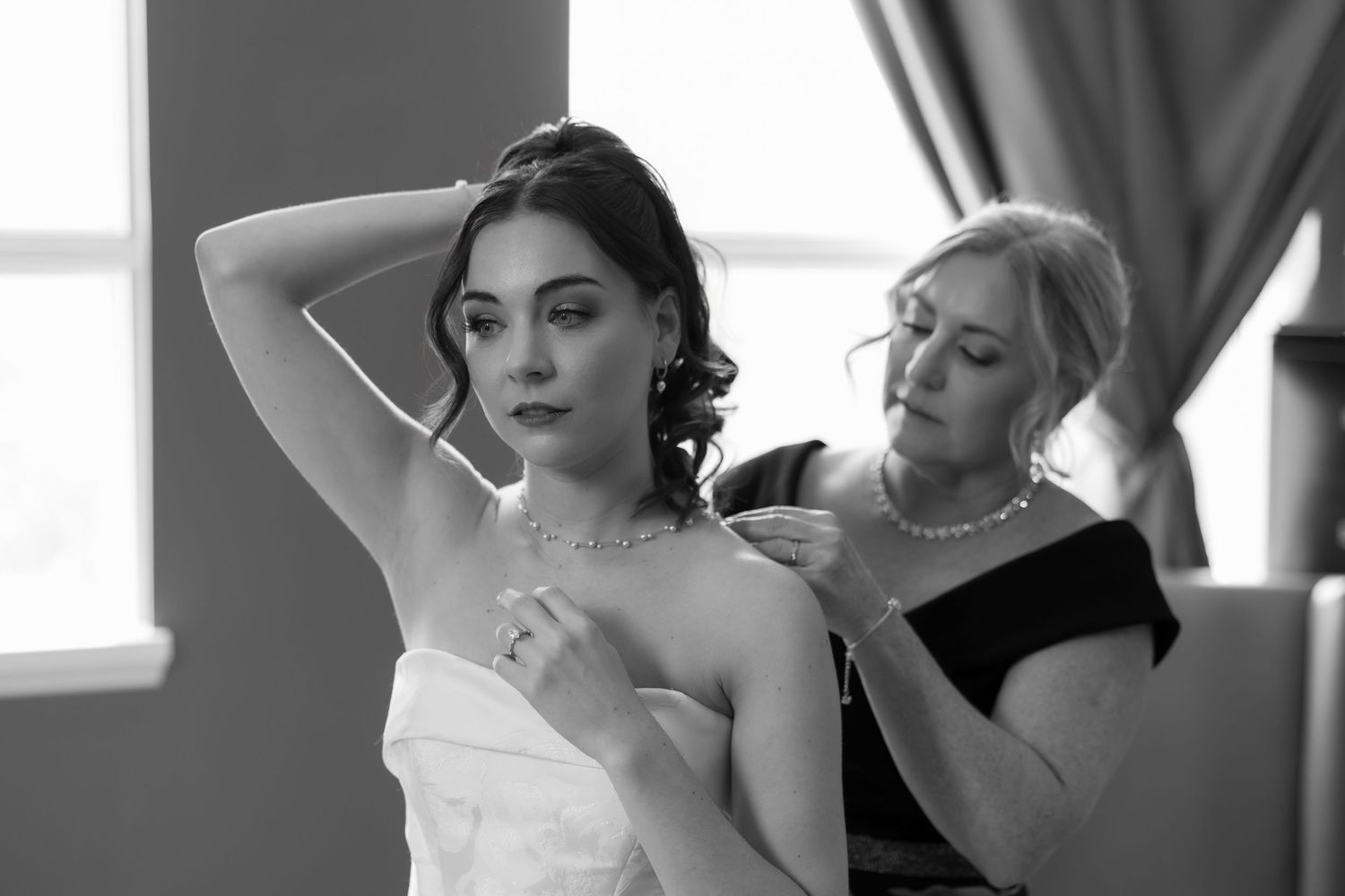 Black and white photo of a bride having her necklace fastened by a woman in a strapless wedding gown.