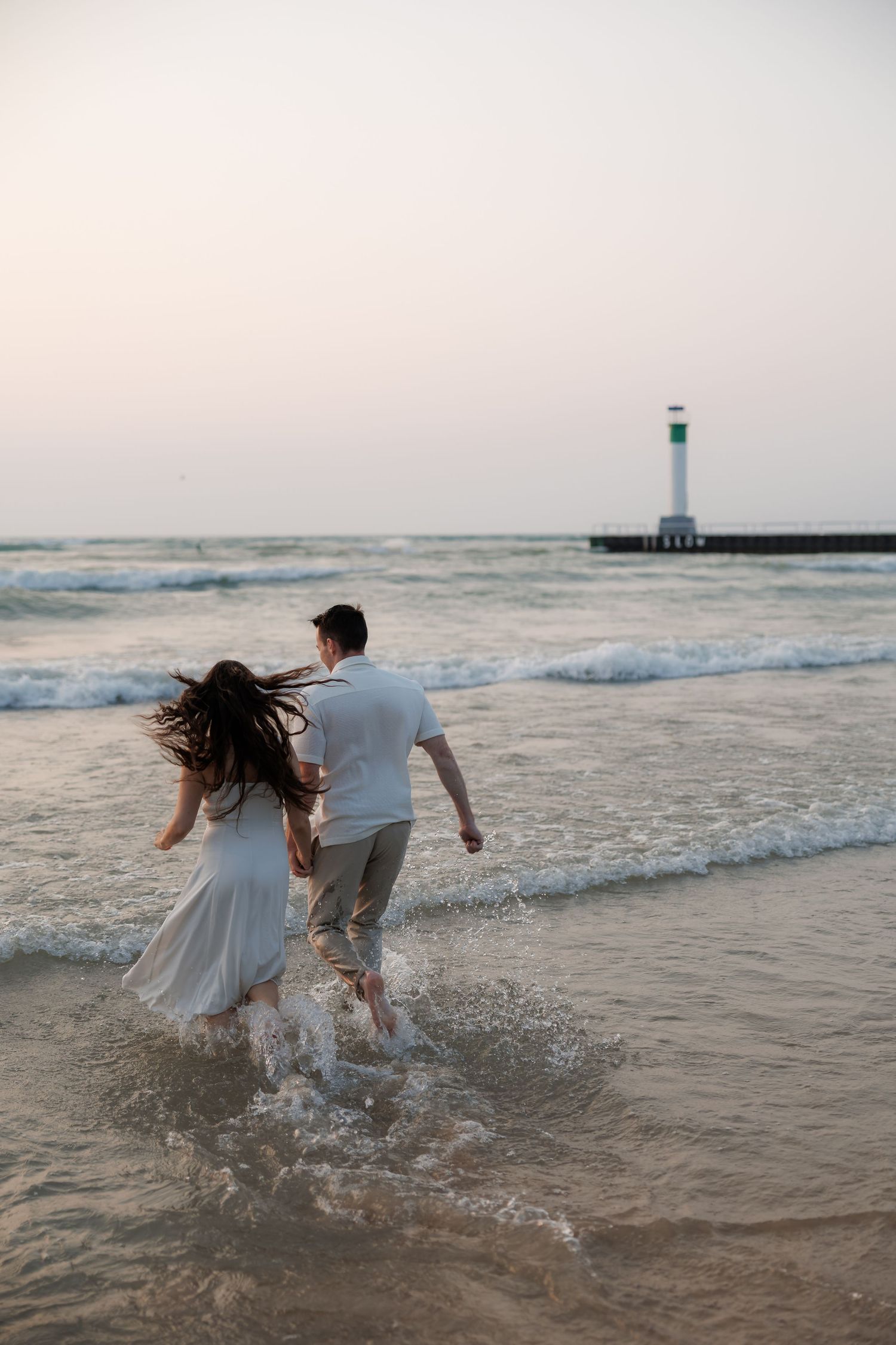 A couple in white clothing playfully splashes in the ocean waves at sunset with a lighthouse visible in the background.