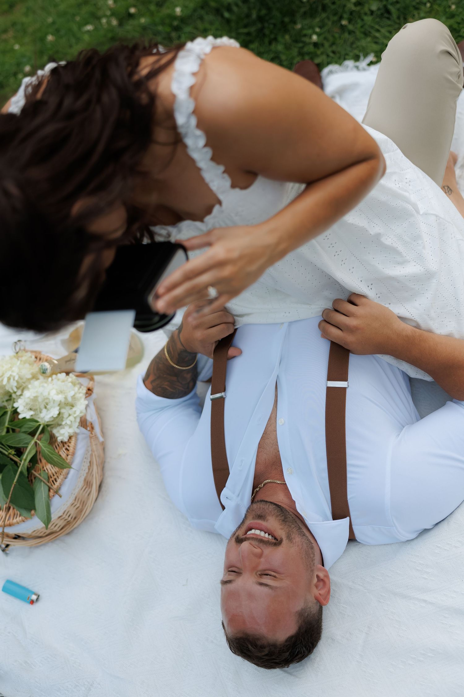 Playful couple in white outfits laughing while lying on picnic blanket outdoors.