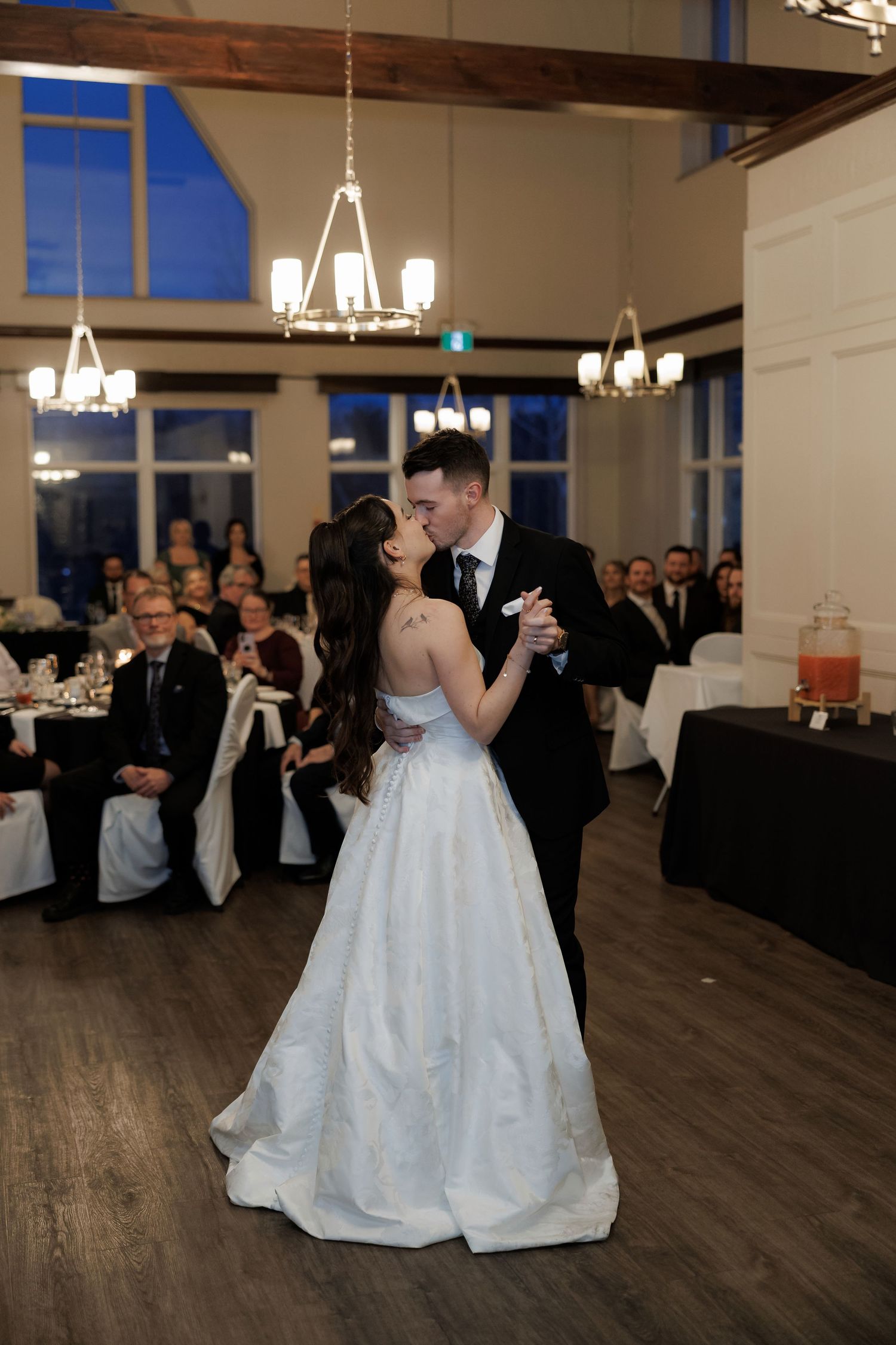 Bride and groom share their first dance in an elegant wedding reception hall with chandeliers and seated guests.