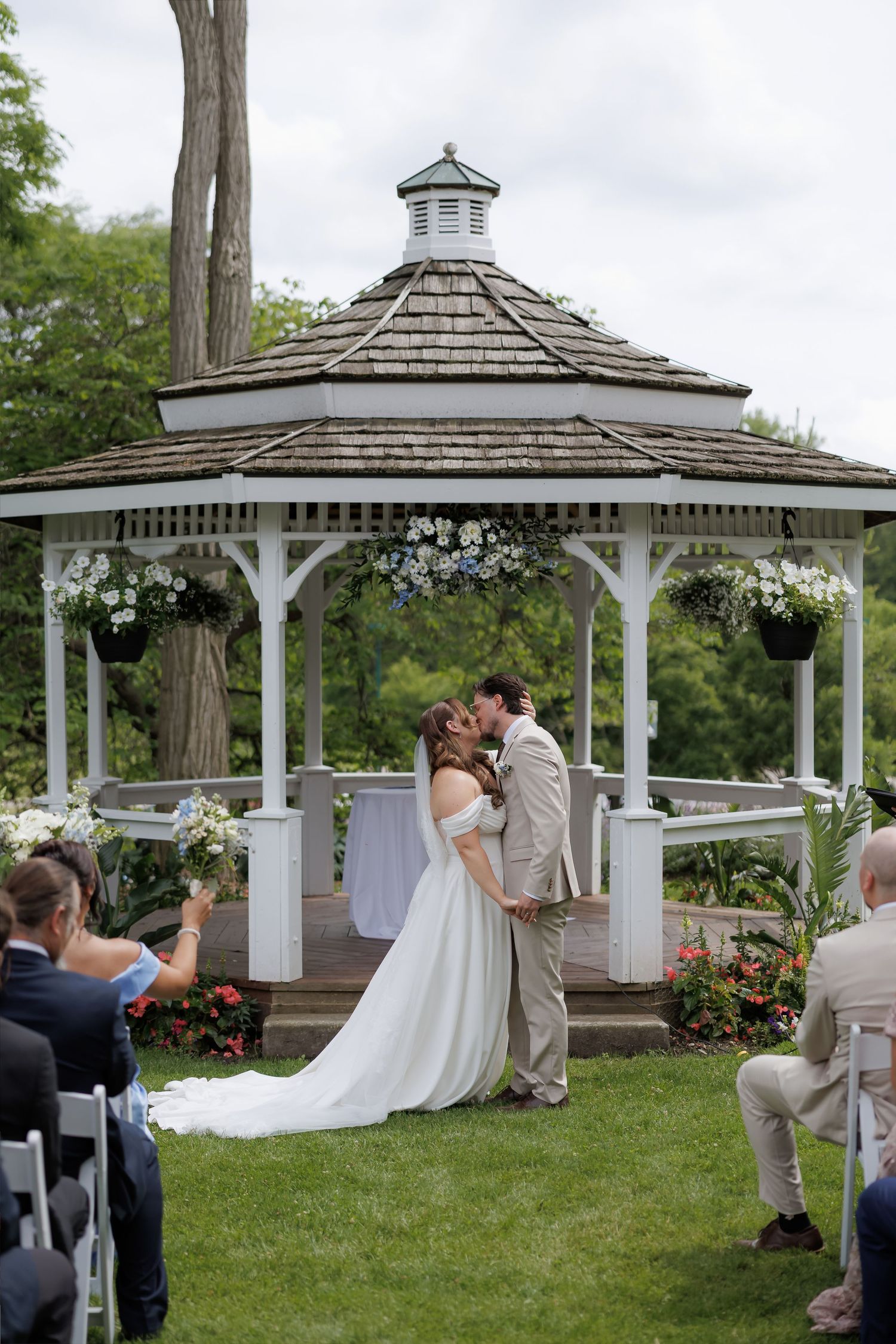 Bride and groom share first kiss under a white floral gazebo during outdoor wedding ceremony with guests seated nearby.