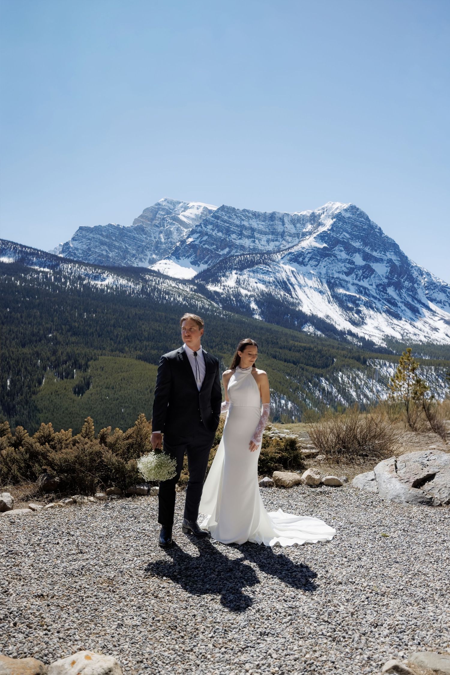Bride and groom pose on rocky terrain with snow-capped mountain backdrop under clear blue sky.