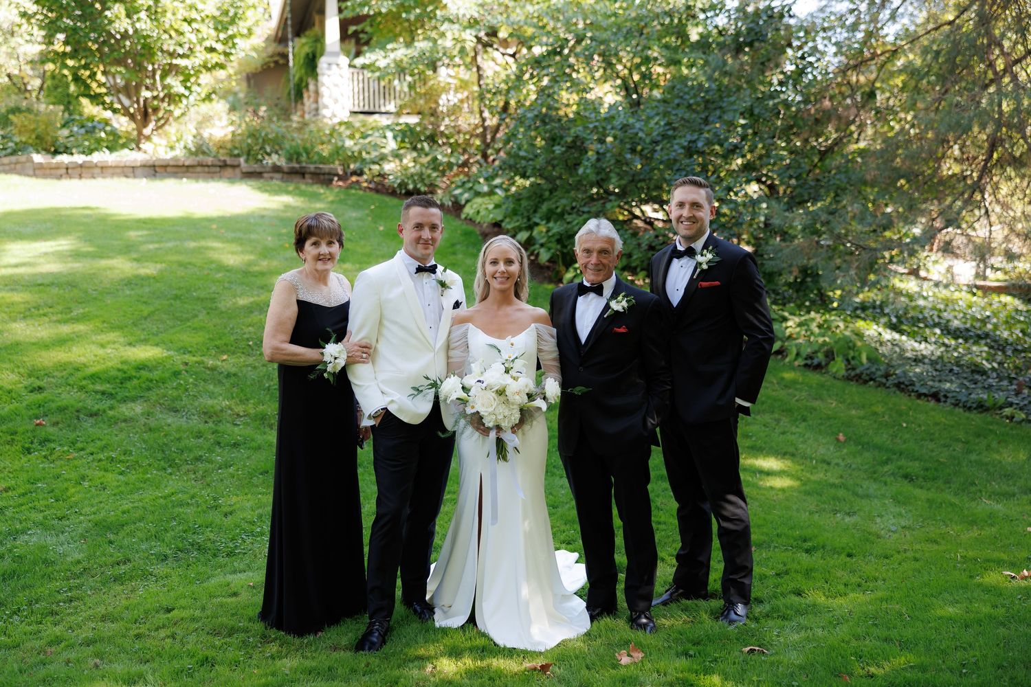 Wedding party in formal attire poses on green lawn with trees in background.