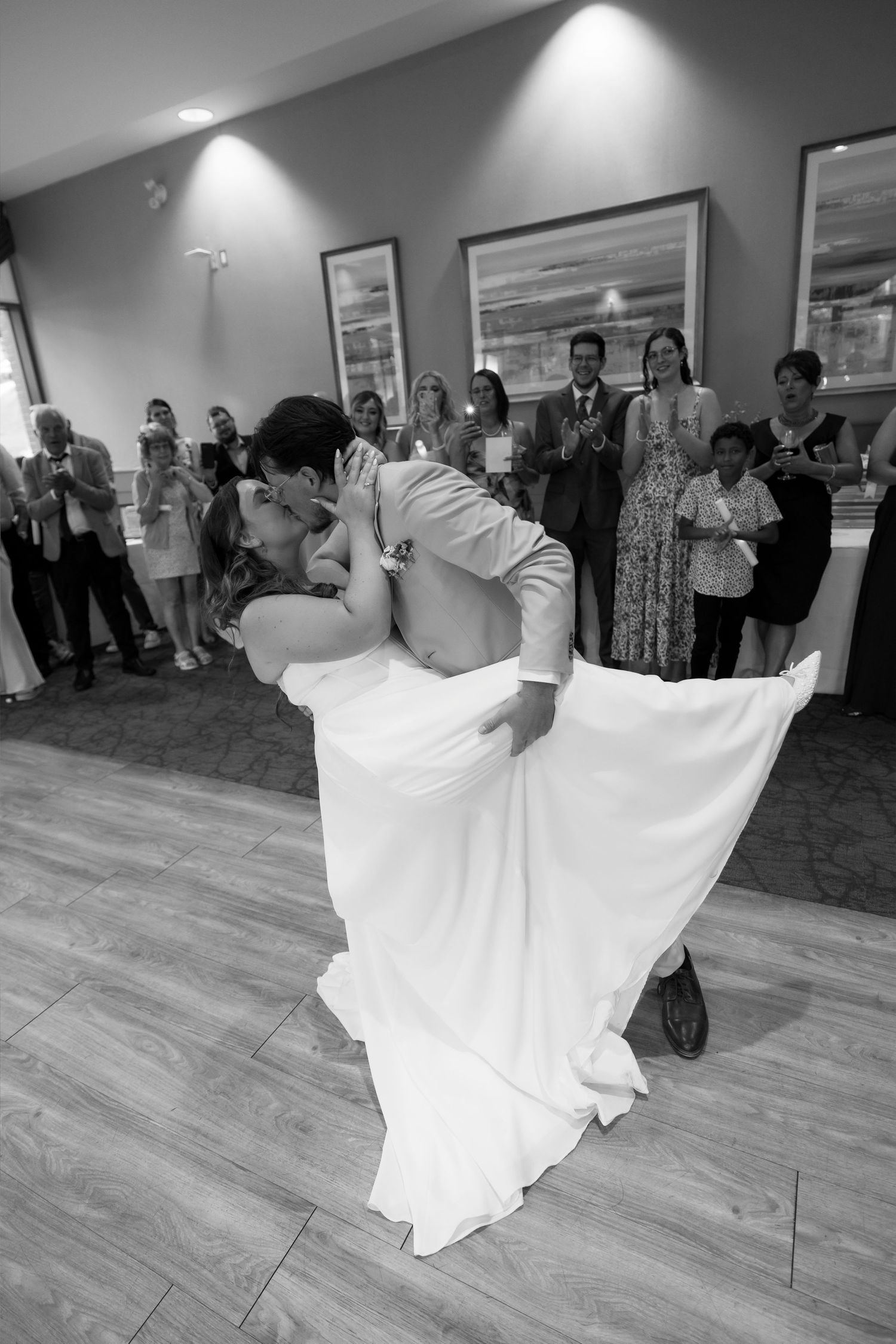 Bride and groom share romantic dip kiss during first dance at wedding reception while guests watch and applaud.