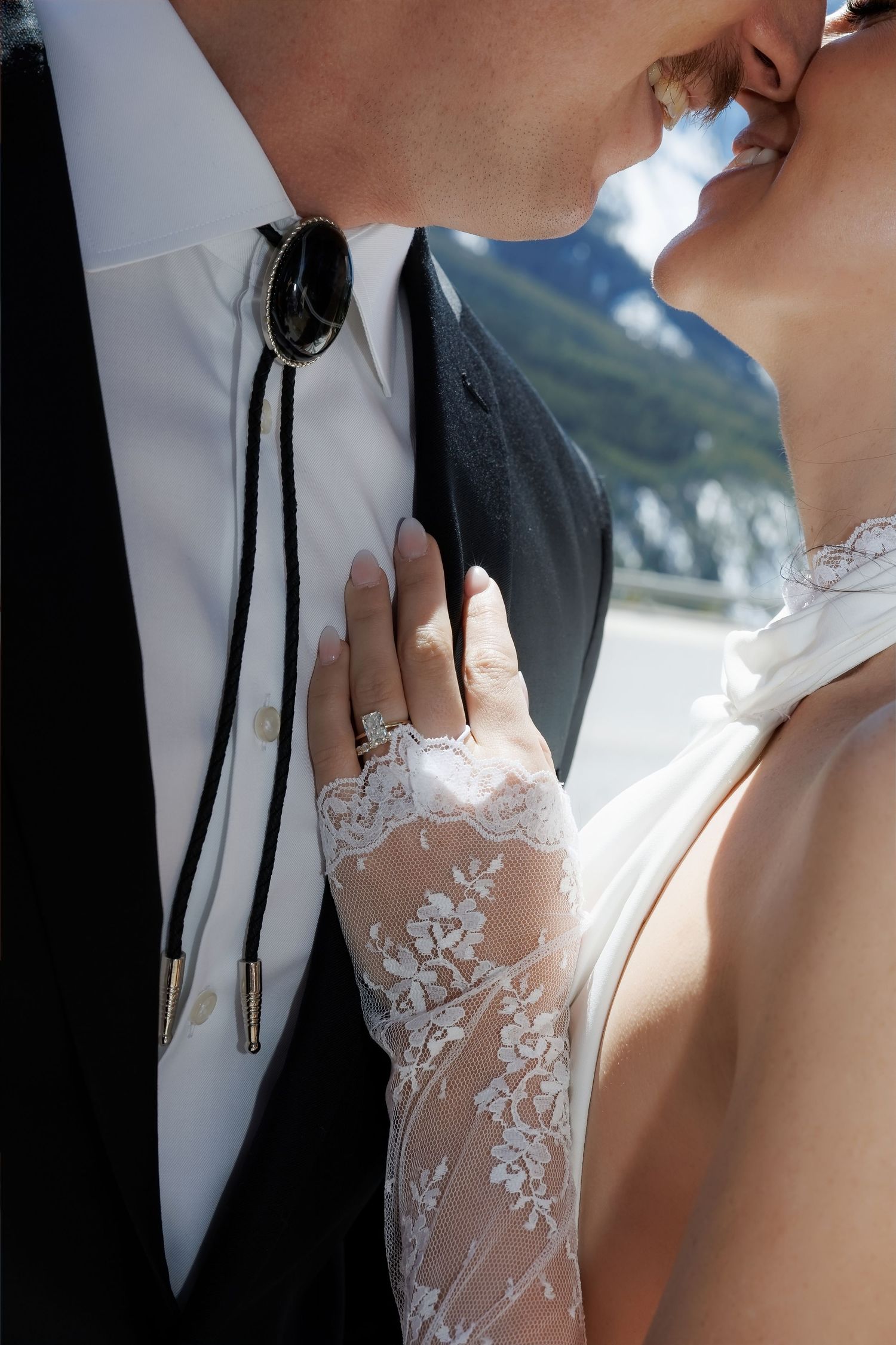 Bride and groom sharing a kiss outdoors, bride wearing lace gloves and wedding ring with mountains in background.