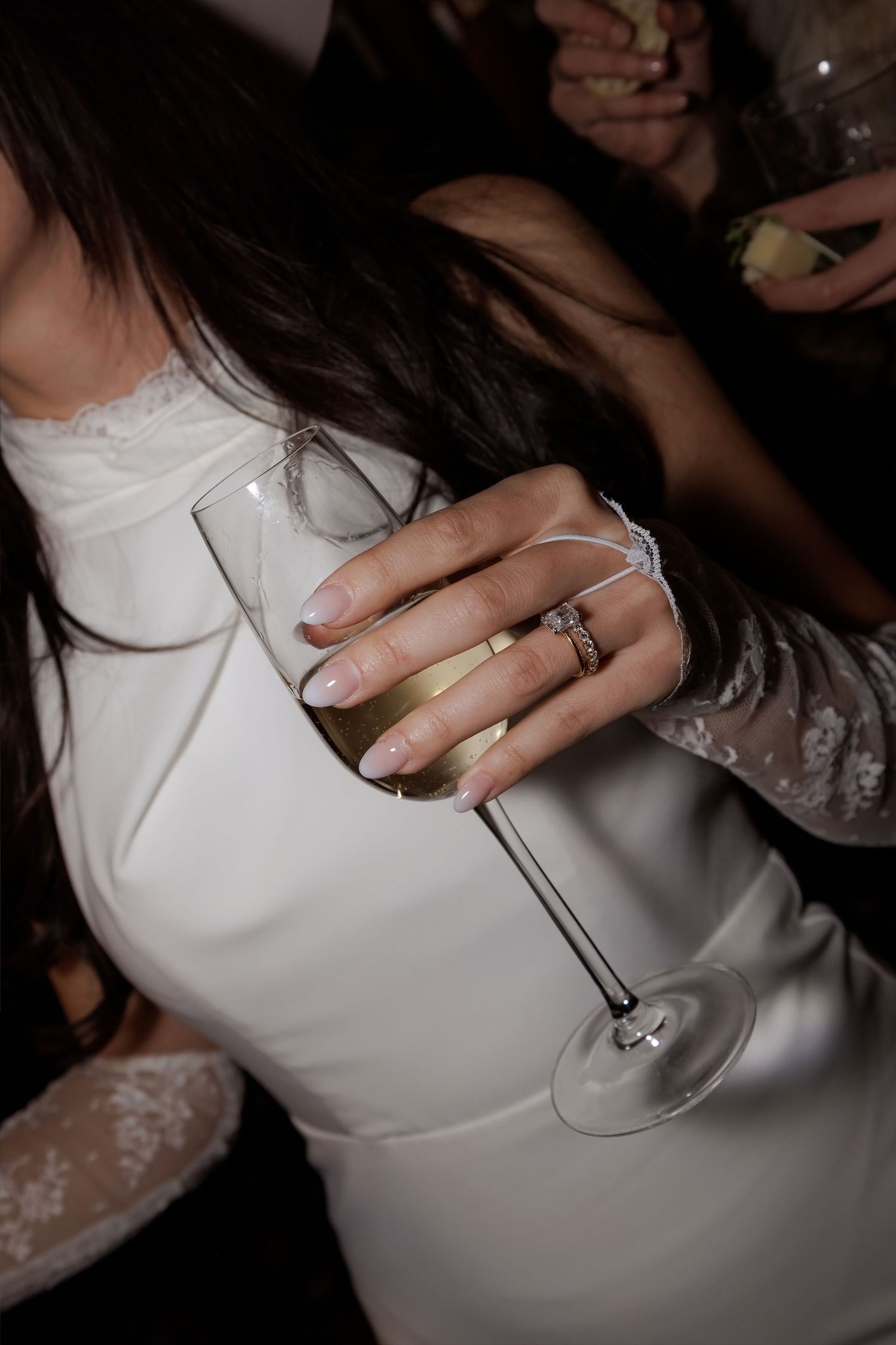 Bride in white lace wedding dress holding champagne flute with diamond engagement ring and wedding band.
