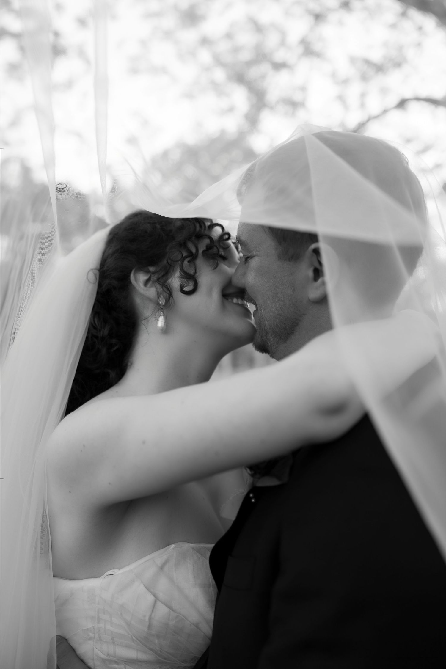 Black and white wedding photo of bride and groom kissing under a veil outdoors.