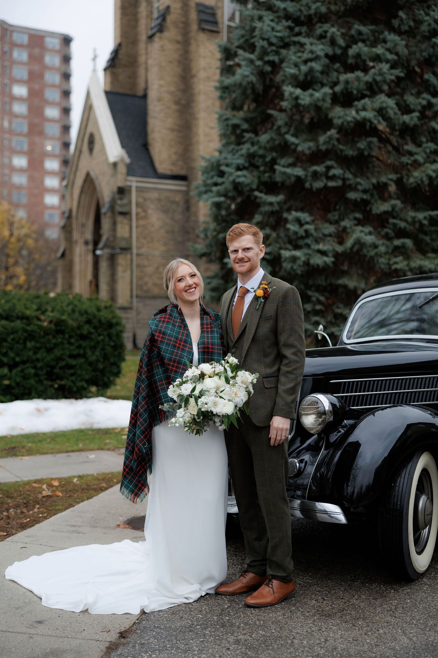 Wedding couple poses with vintage black car outside brick church in winter.