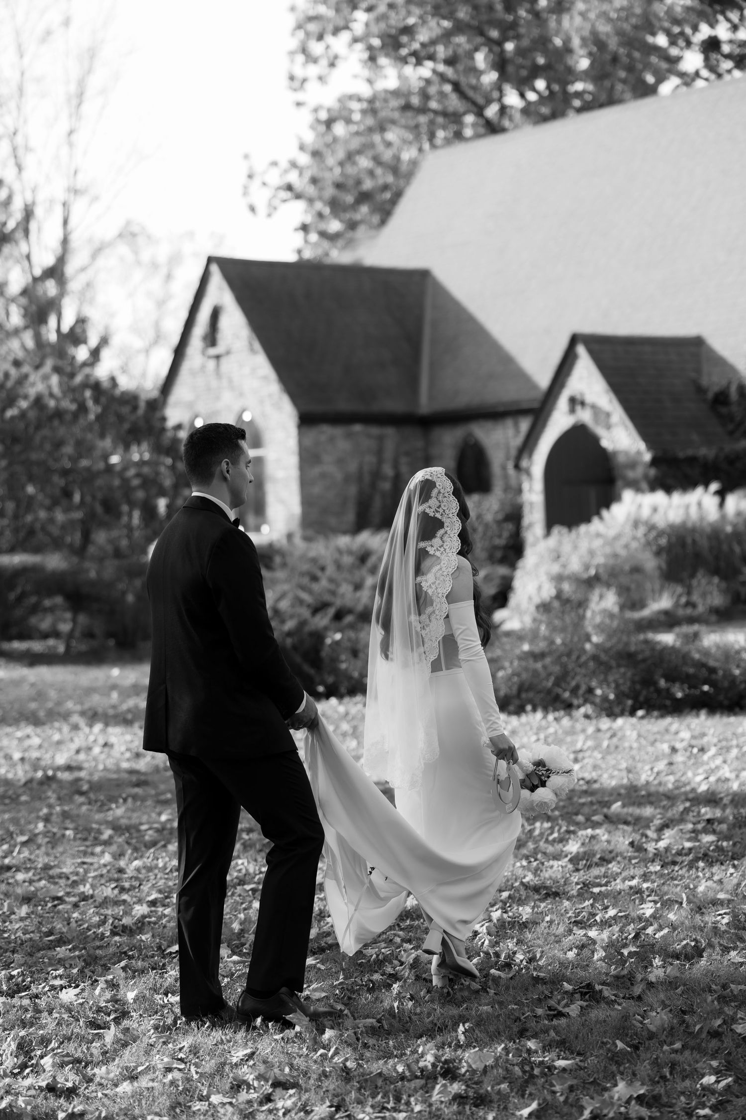 Black and white photo of newlyweds walking together on leaf-covered ground near historic buildings.