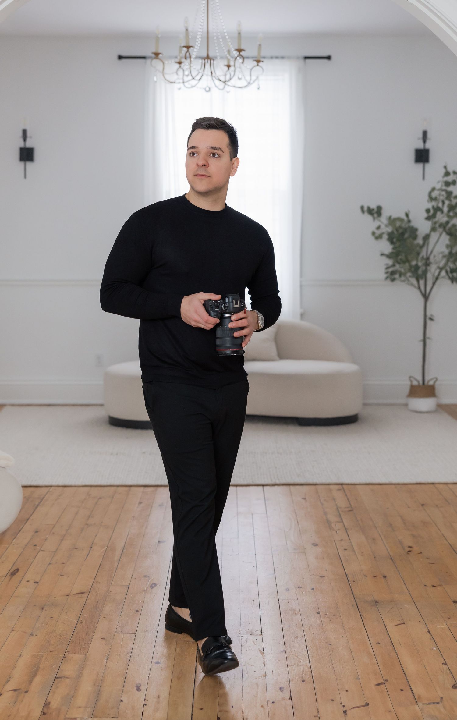 Person in black clothing walking across a bright white room with wooden floors and chandelier.