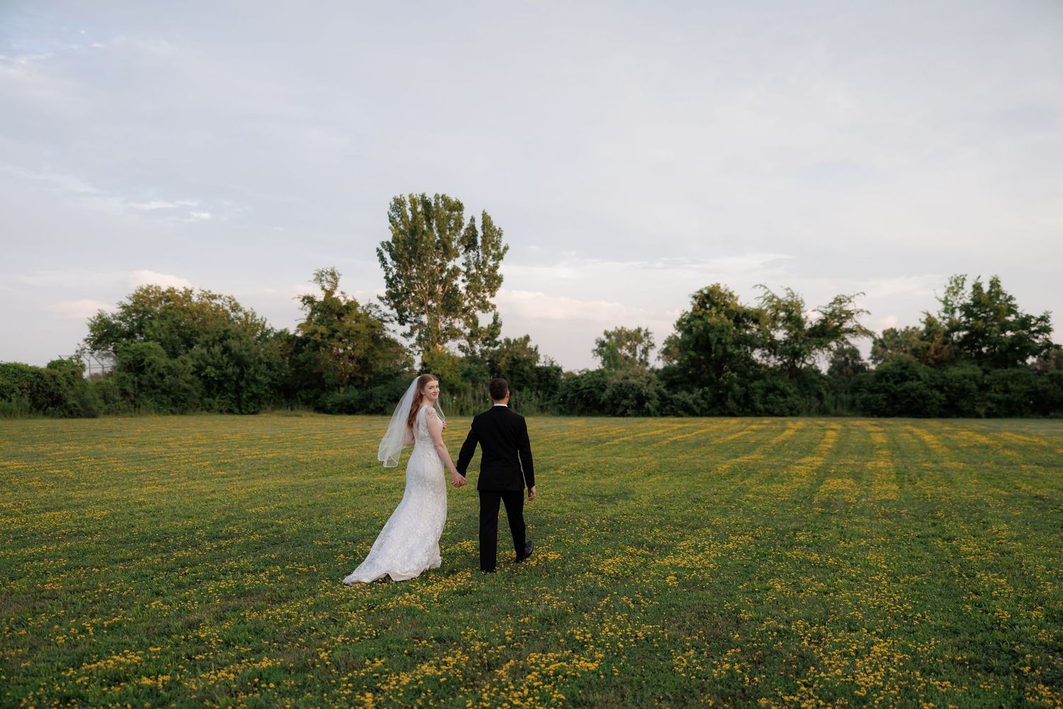 Newlyweds walk hand in hand through a field of yellow wildflowers at sunset.