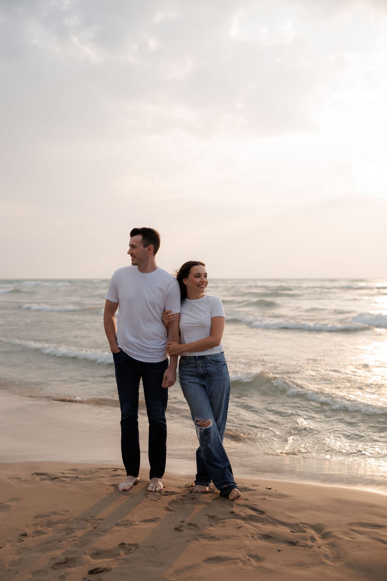 A couple walks together along a sandy beach at sunset with waves gently rolling onto the shore.
