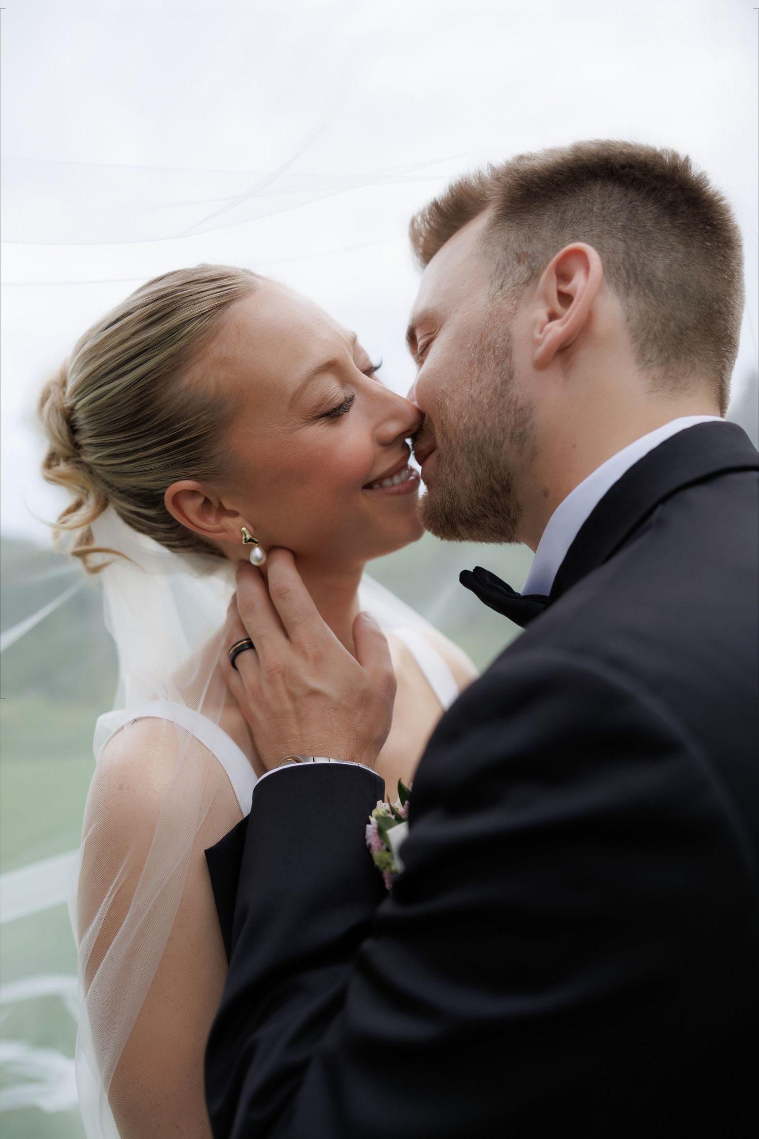Bride and groom sharing a romantic almost-kiss under a flowing veil, groom tenderly holding her face.